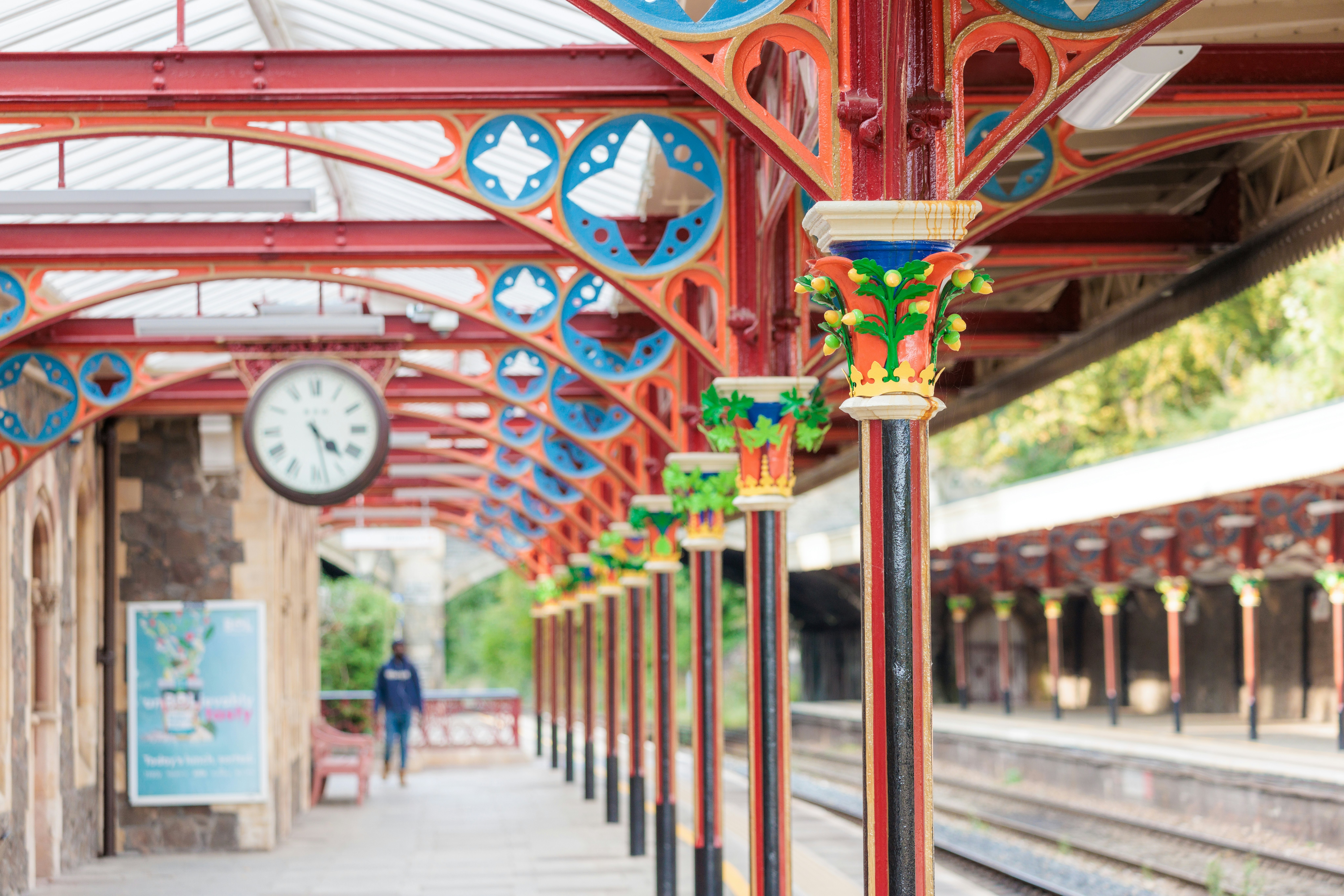 A train platform with red wrought-iron ceiling supports and pillars. There are colorful iron flowers and crown-like decorations on each pillar.