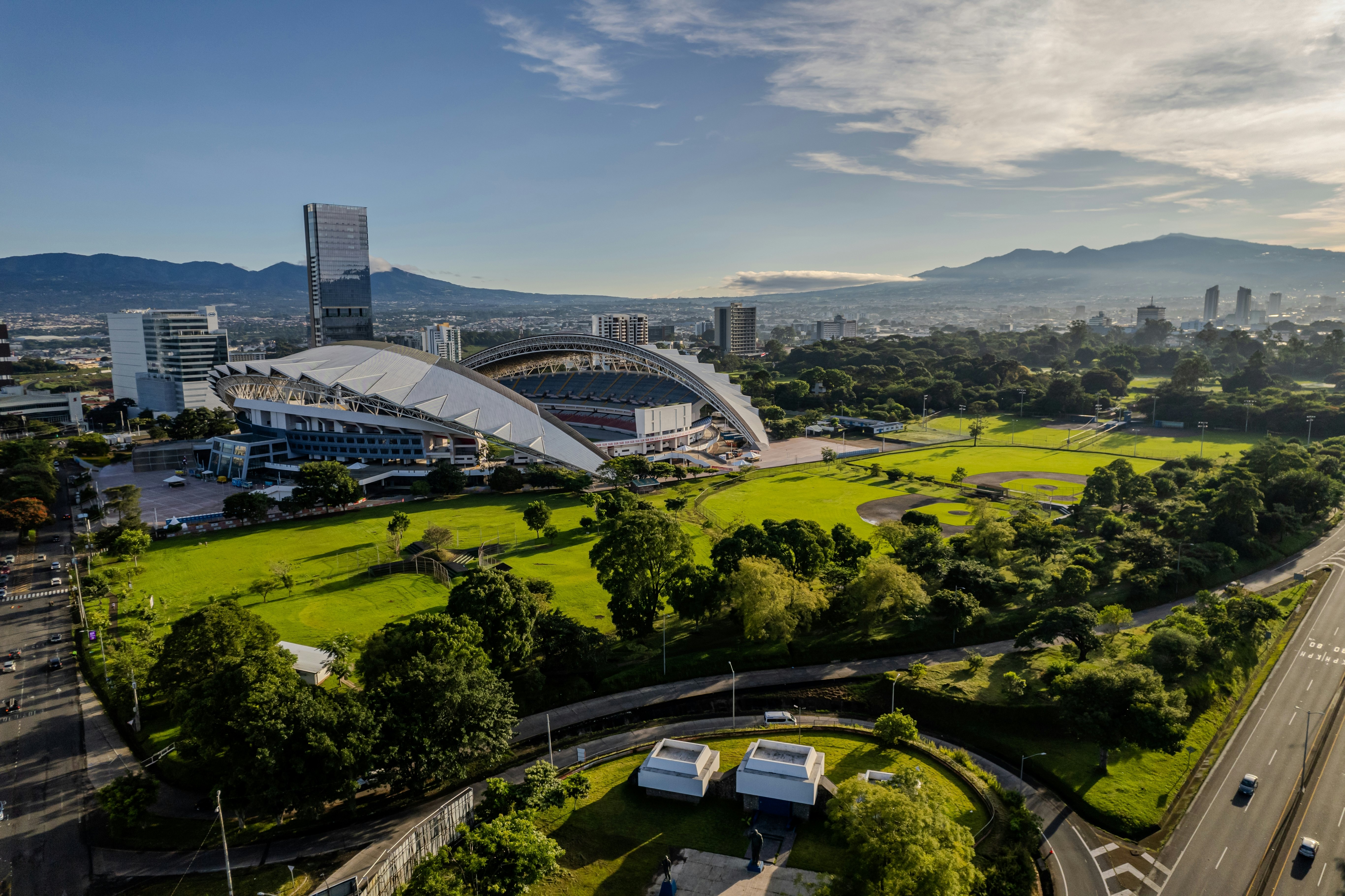 An wide shot of a green park surrounding a stadium in a busy city.