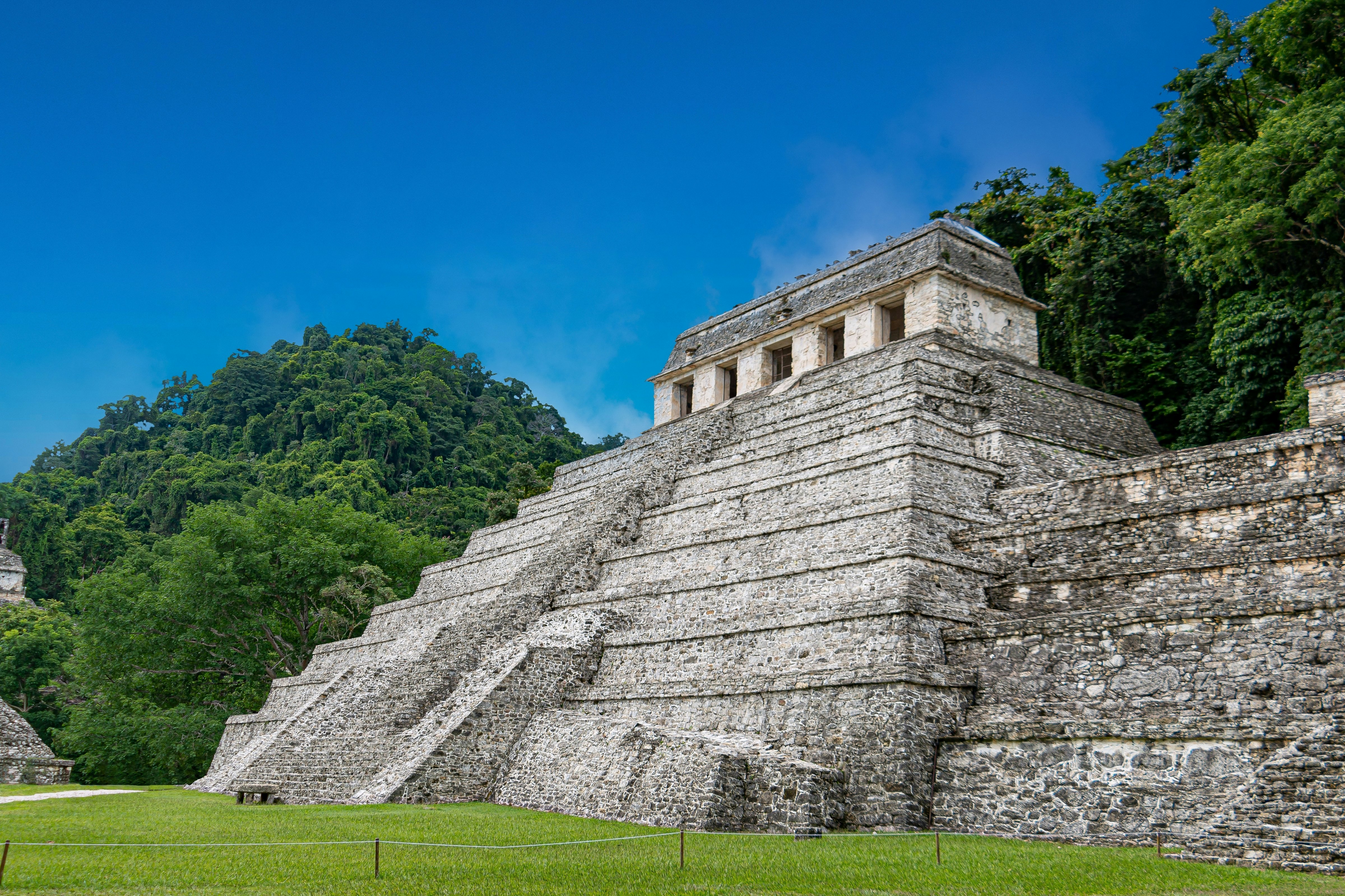 Ancient stepped ruins with green grass in front and trees in the background under a blue sky.