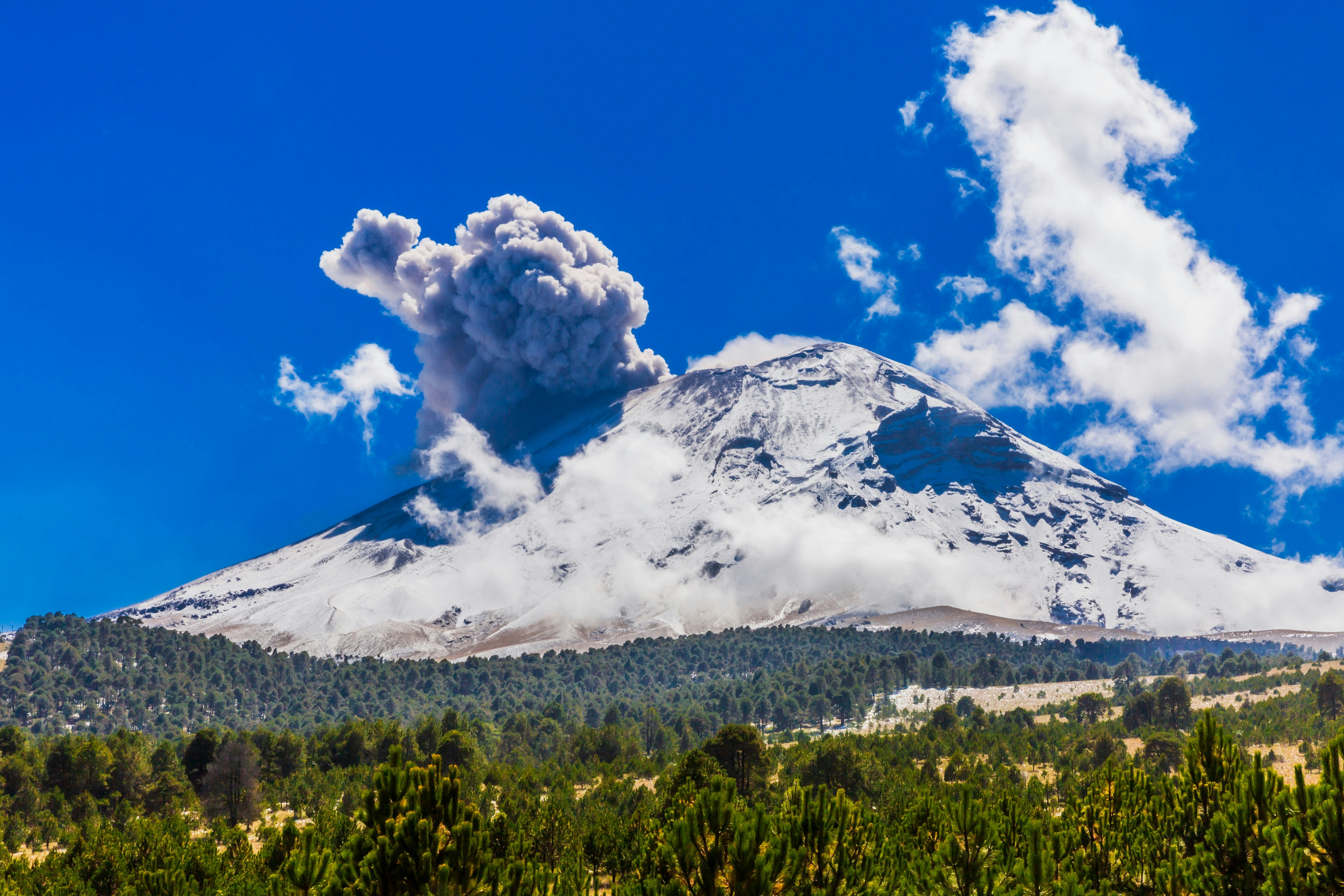 A wide view of a volcano covered with snow and releasing smoke on a sunny day.