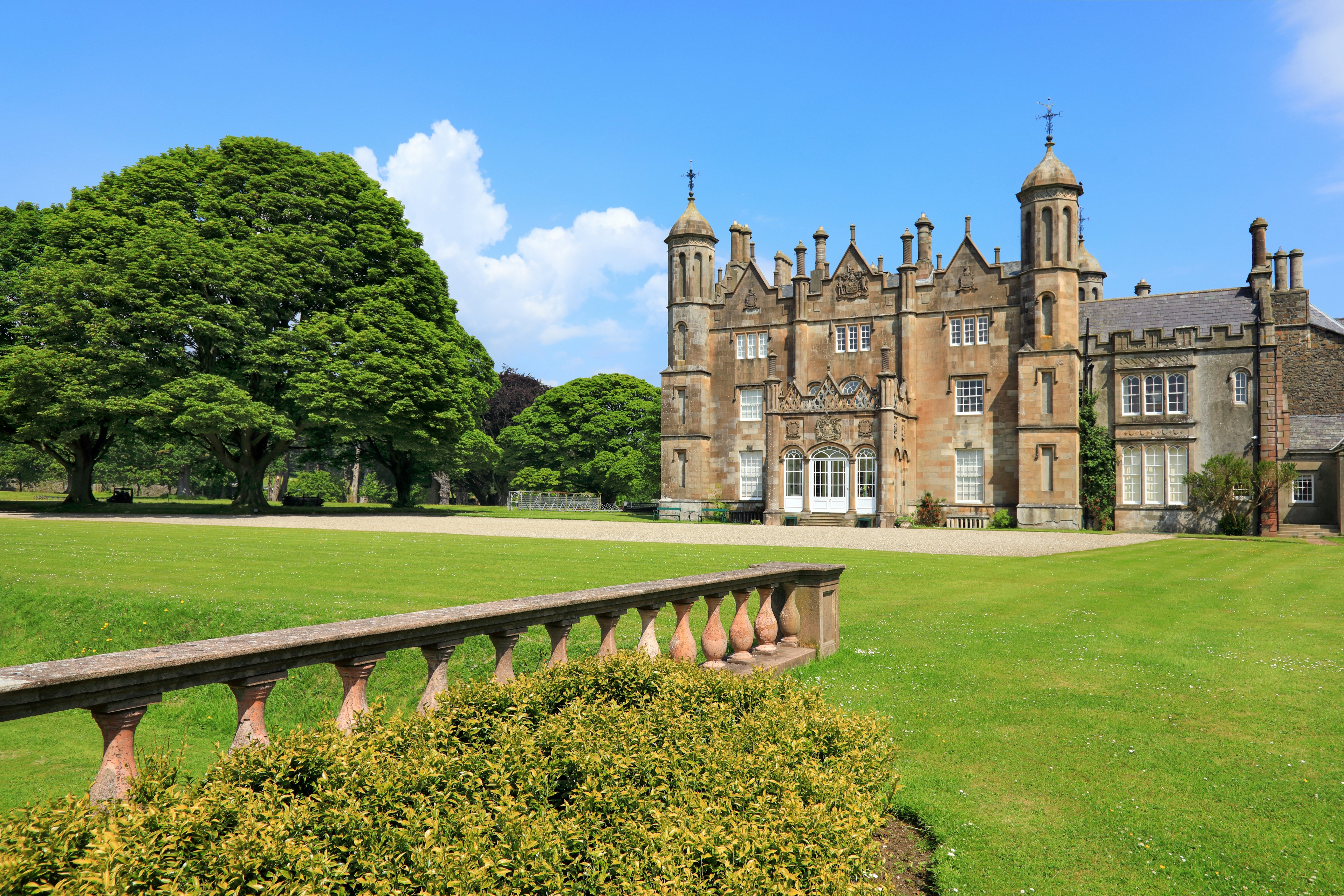 A stone balustrade is seen in a garden, with an ornate manor house across a well-mowed lawn.
