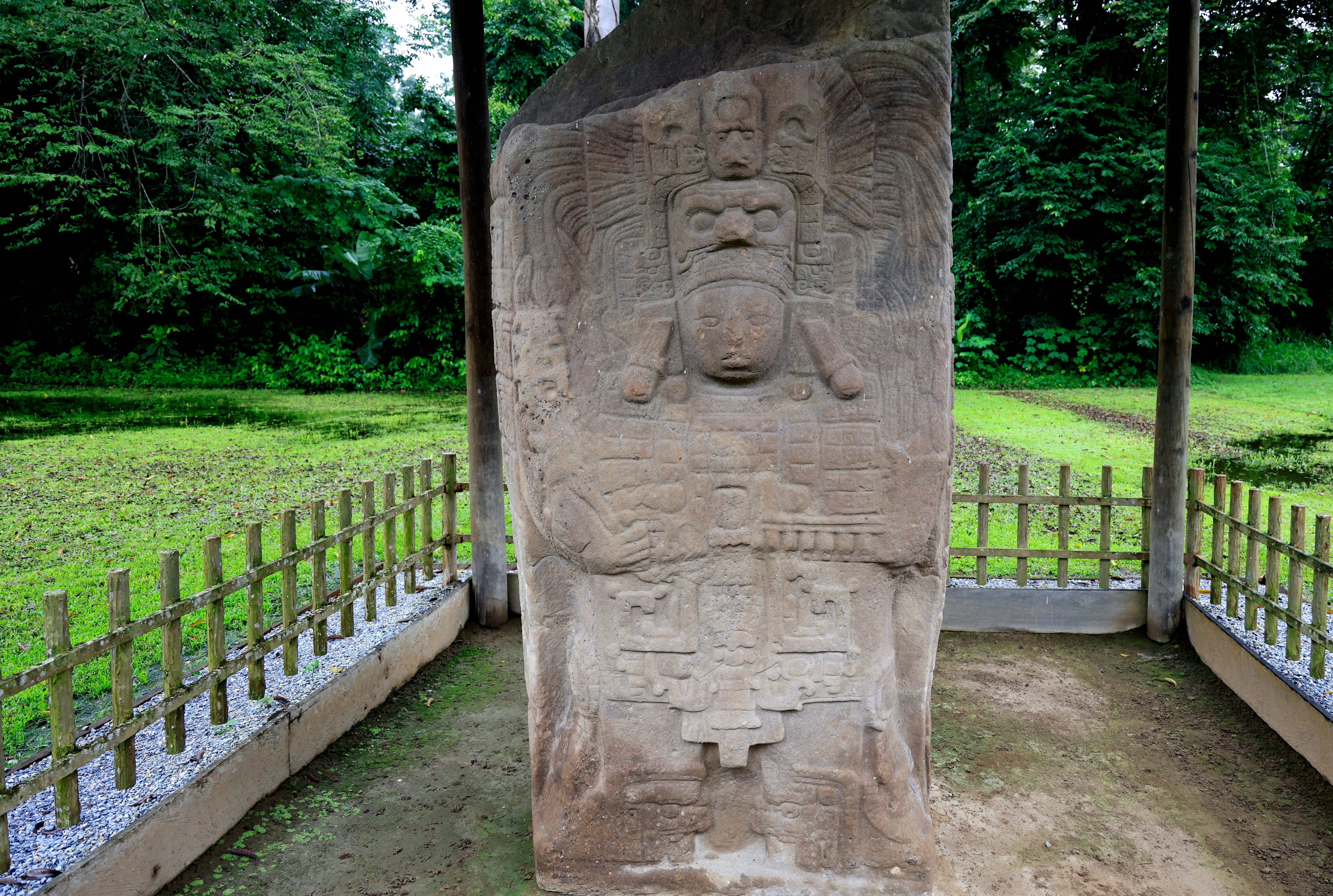 A stone stele on display outdoors behind a fence is carved with stylized faces and other motifs.