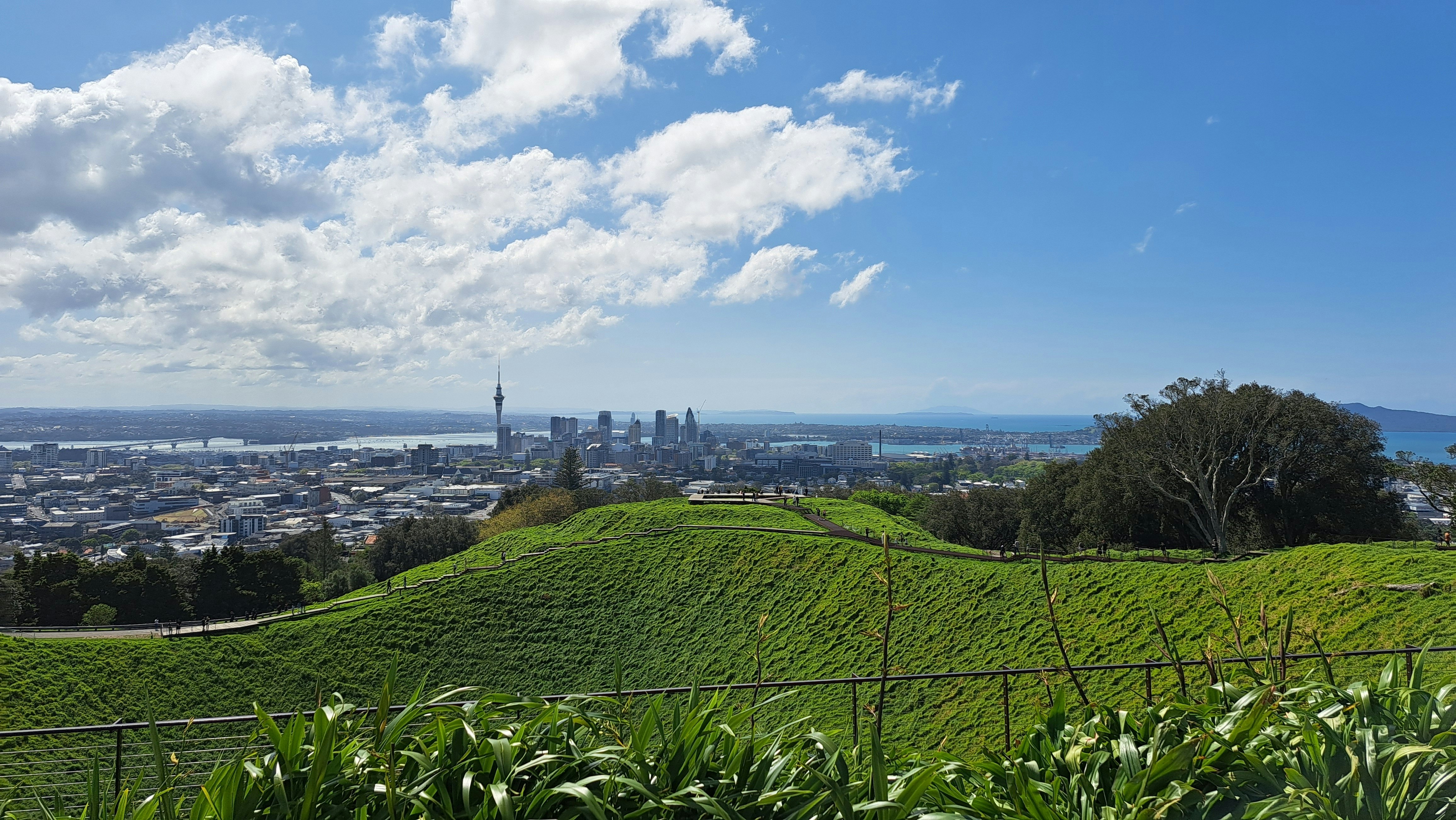 A green hill with a path at the top is in the foreground and the skyline of Auckland, New Zealand, is in the background under blue sky with some clouds.