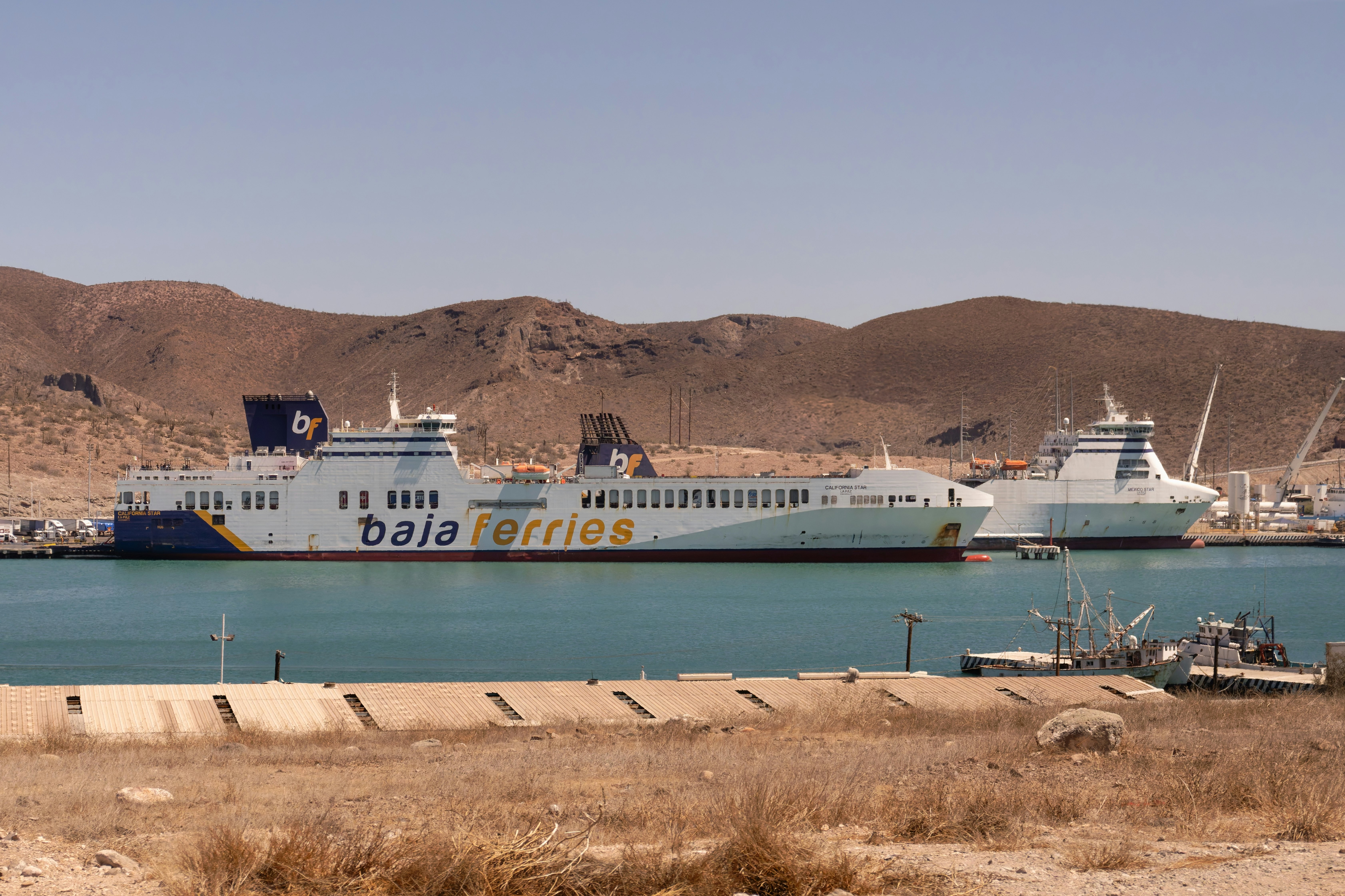 A large passenger ferry is moored in a port. The hills of a desert landscape are seen in the distance.