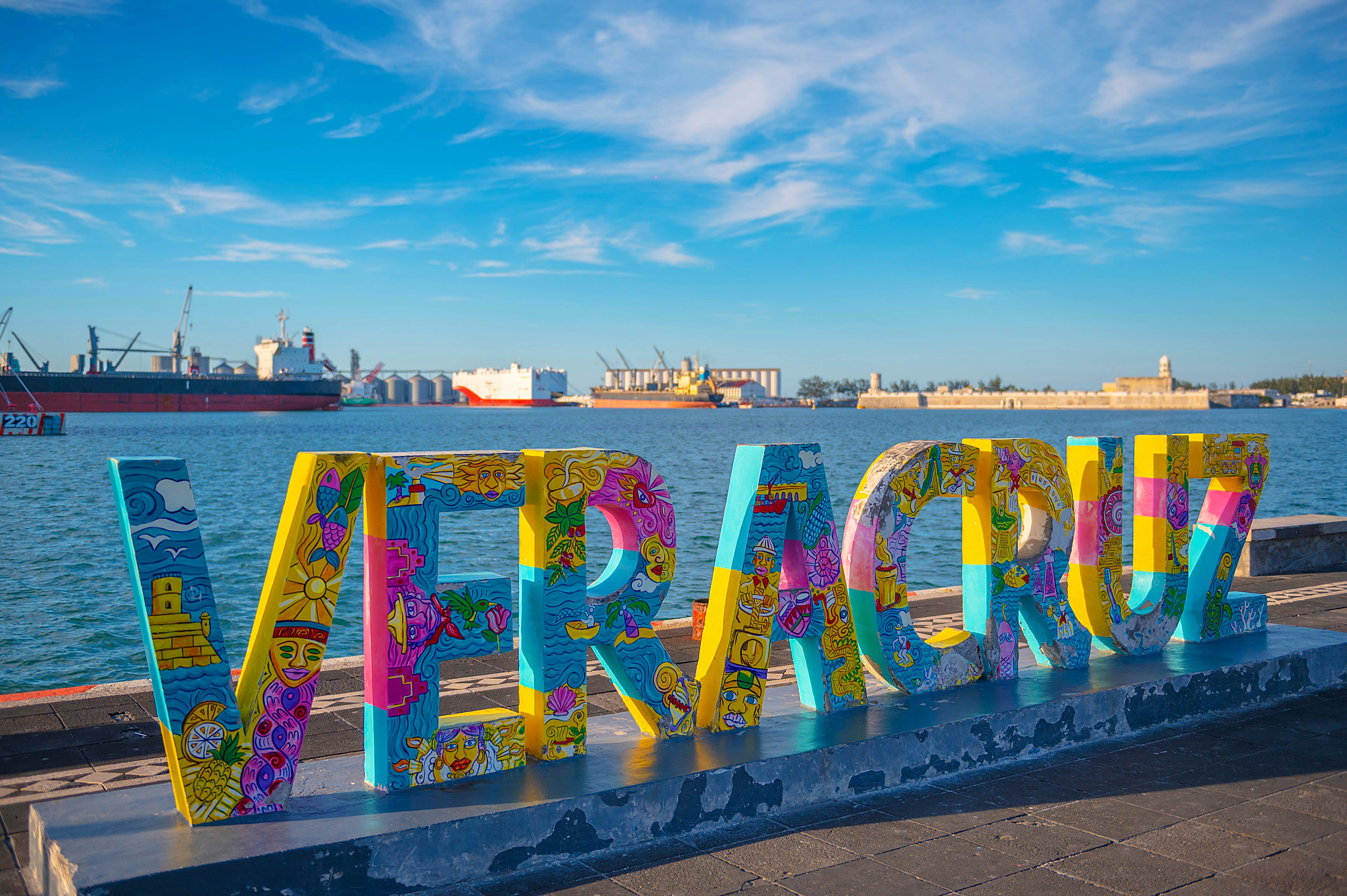 A colorfully painted sign of tall letters spelling "Veracruz" on a waterfront promenade overlooking a port wih cargo ships in the background on a sunny day.