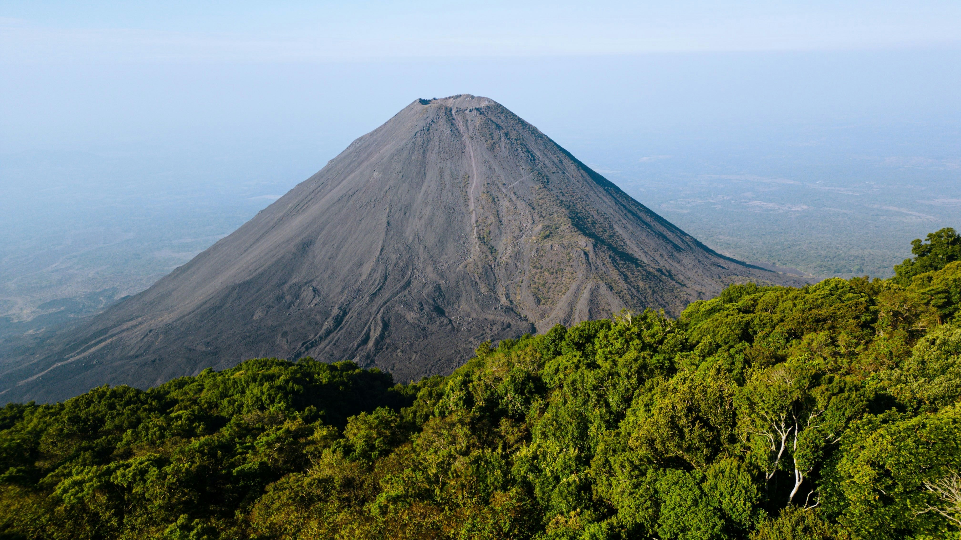 The gray dusty surface of the cone of a volcano surrounded by dense jungle.