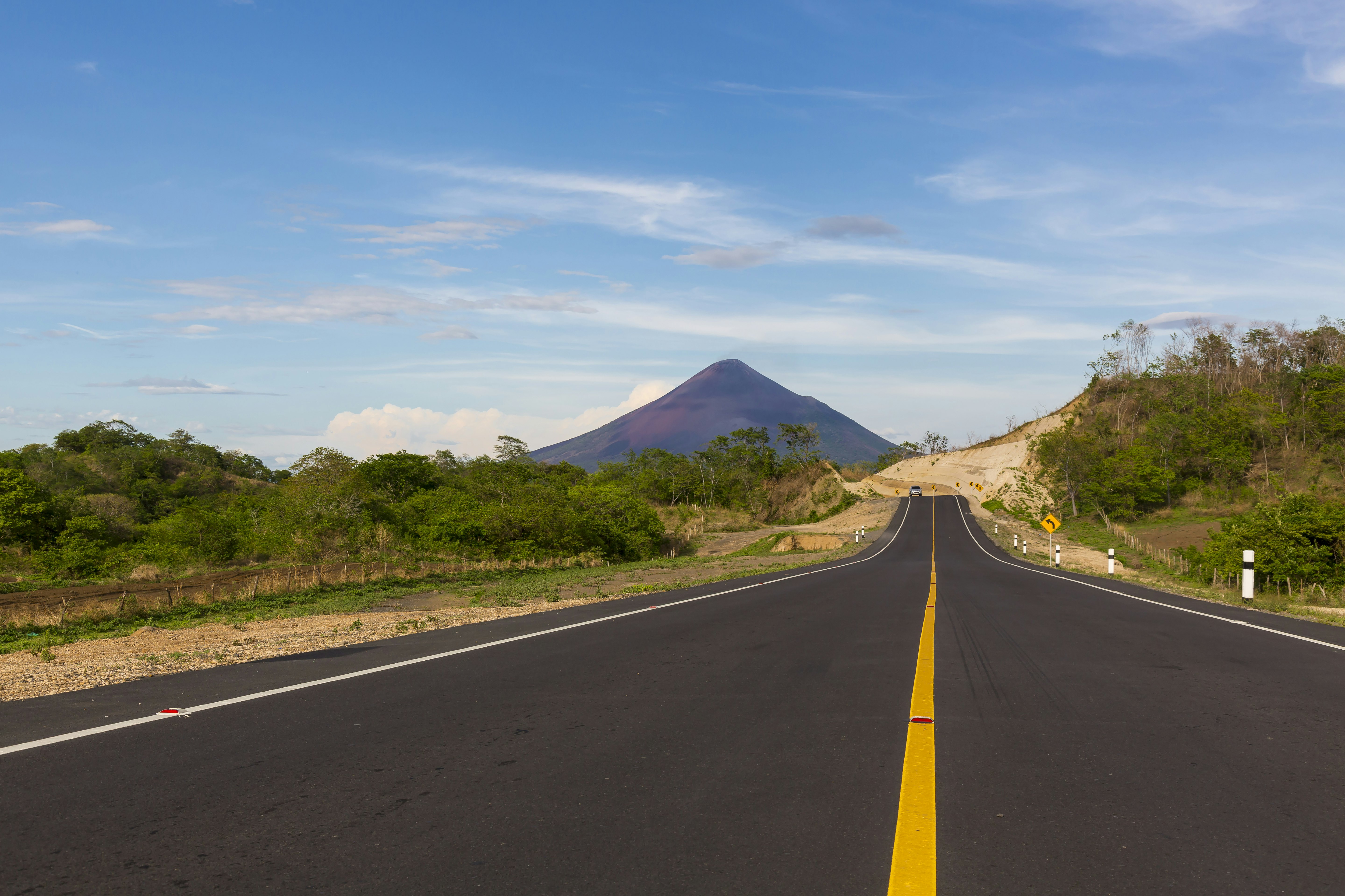 A long, smooth, asphalt road leads through countryside towards a pointed volcano.