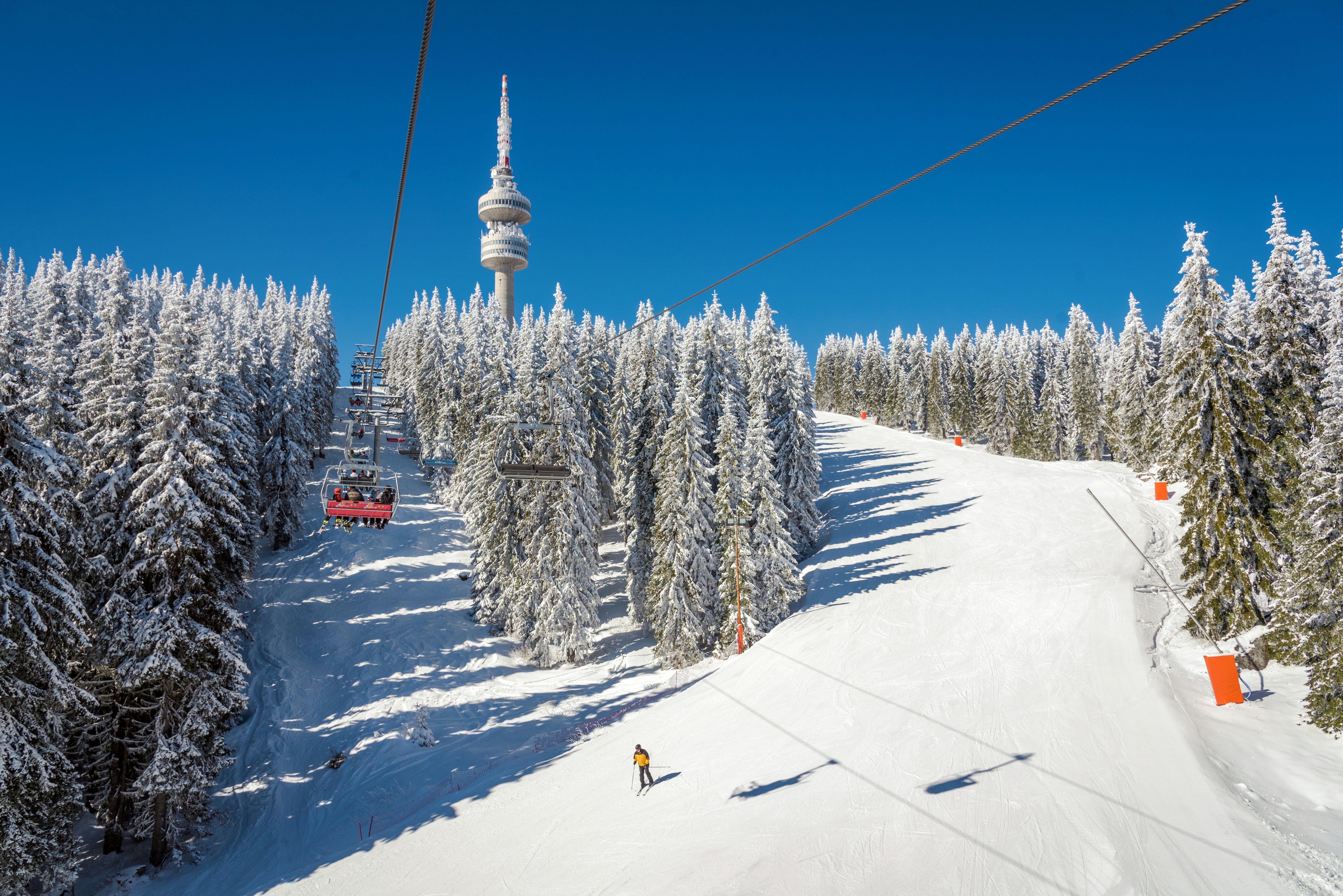 Between tall, snow-covered evergreen trees, the wires of a ski lift suspend people in chairs over one open lane of snow, and on the other side of the trees, another open lane has one skier going downhill. The sky is blue and cloudless, and there is a tower in the middle of the frame.