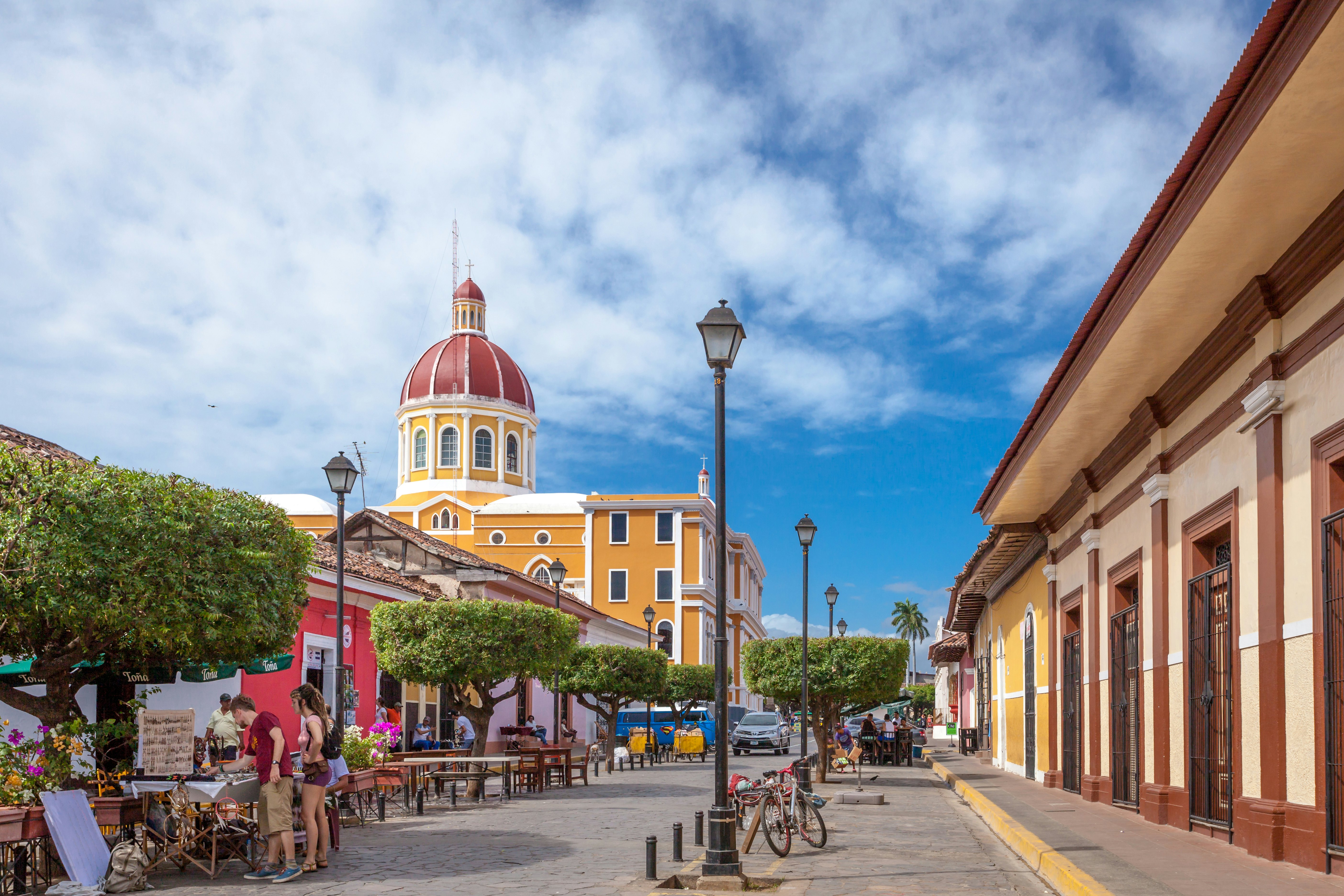 A domed cathdral near a street with colonial-style buildings. Bikes are parked nearby and people browse at a small market stall.