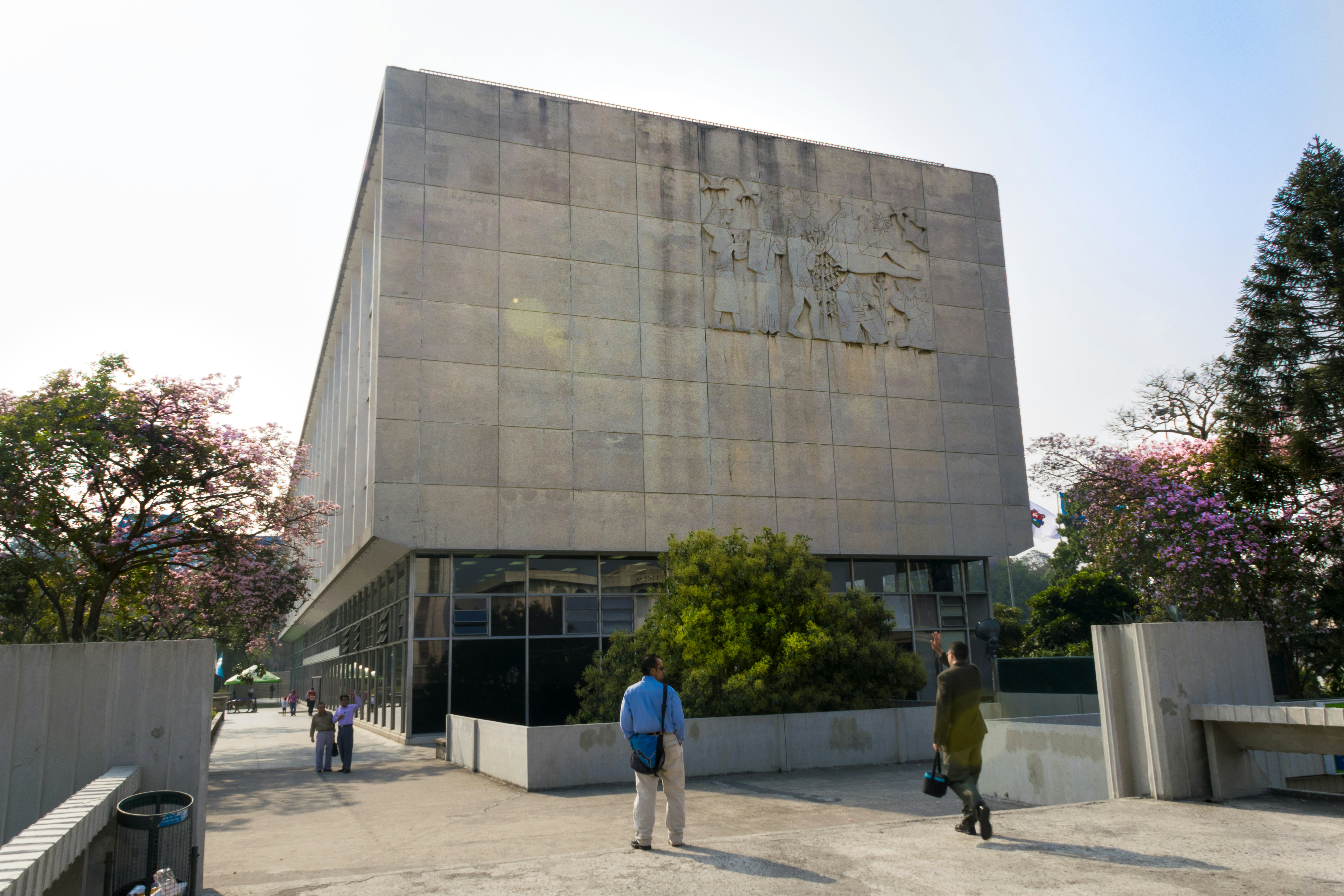 People walk in a city outside a modern building with a relief sculpture on one side.