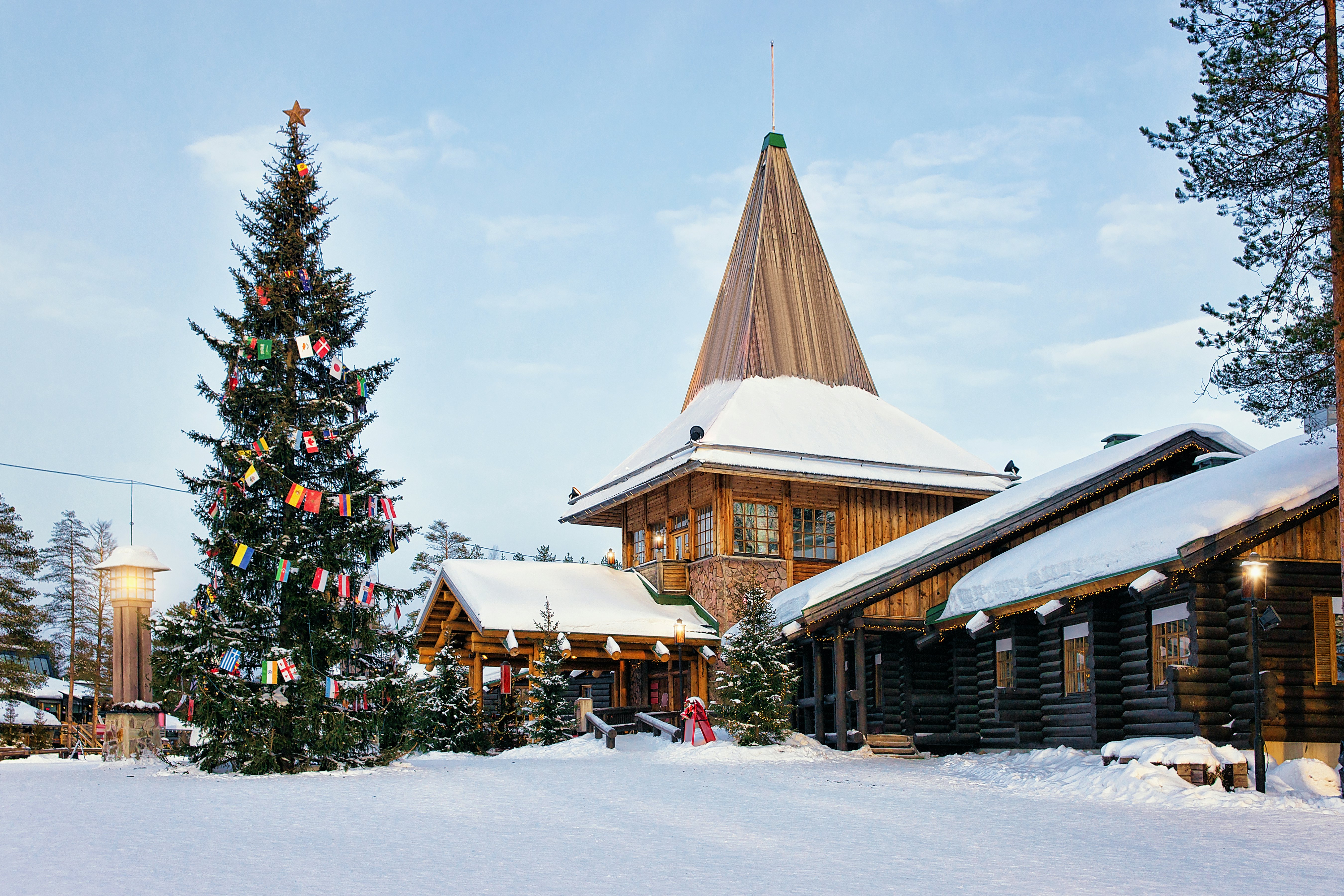A stone and wood building with a steep peak; there is snow on the roof and ground and an evergreen tree decorated with flags.