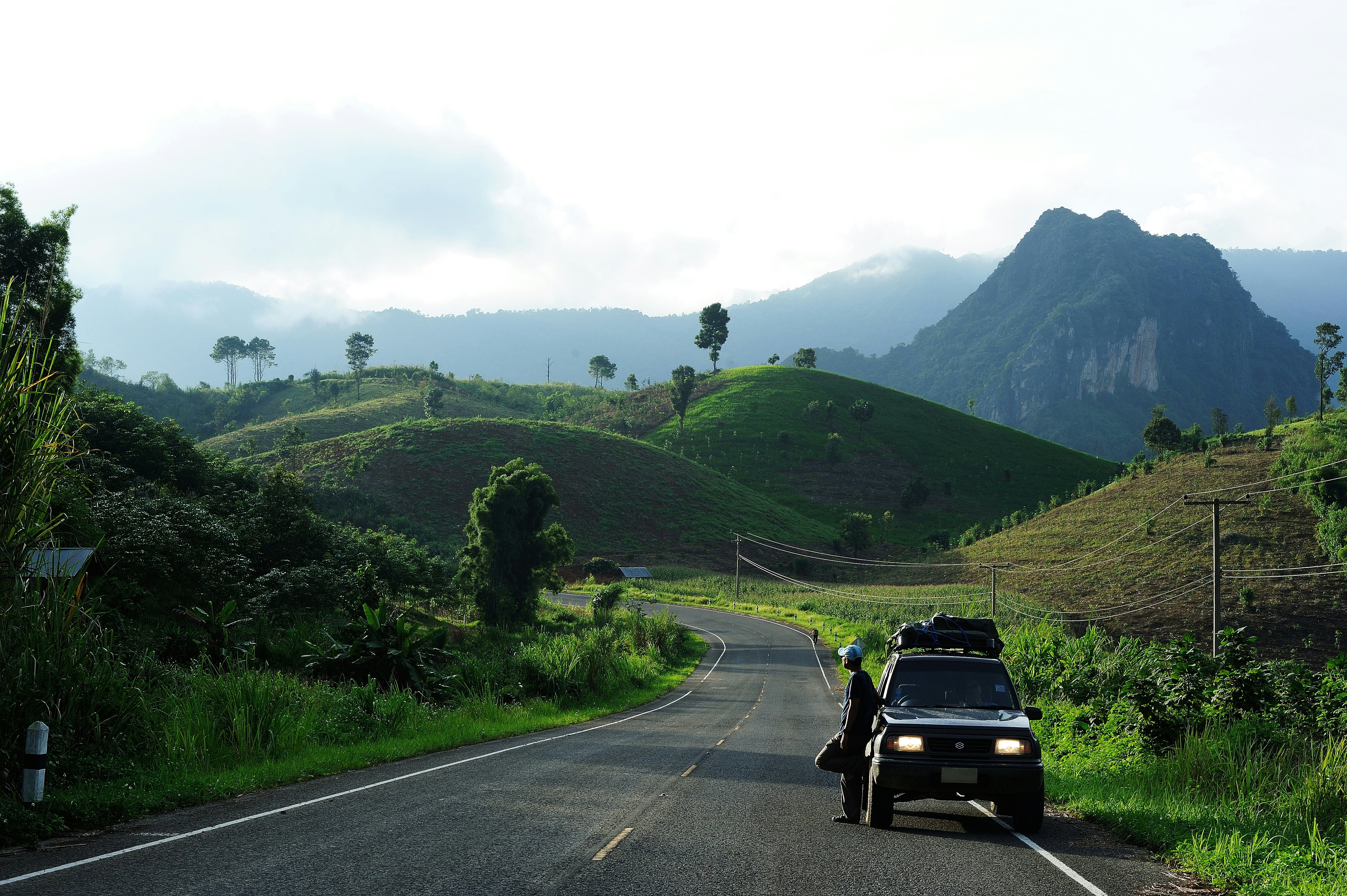 A man stands next to a jeep pulled over on a road through a lush landscape with misty mountains in the distance.