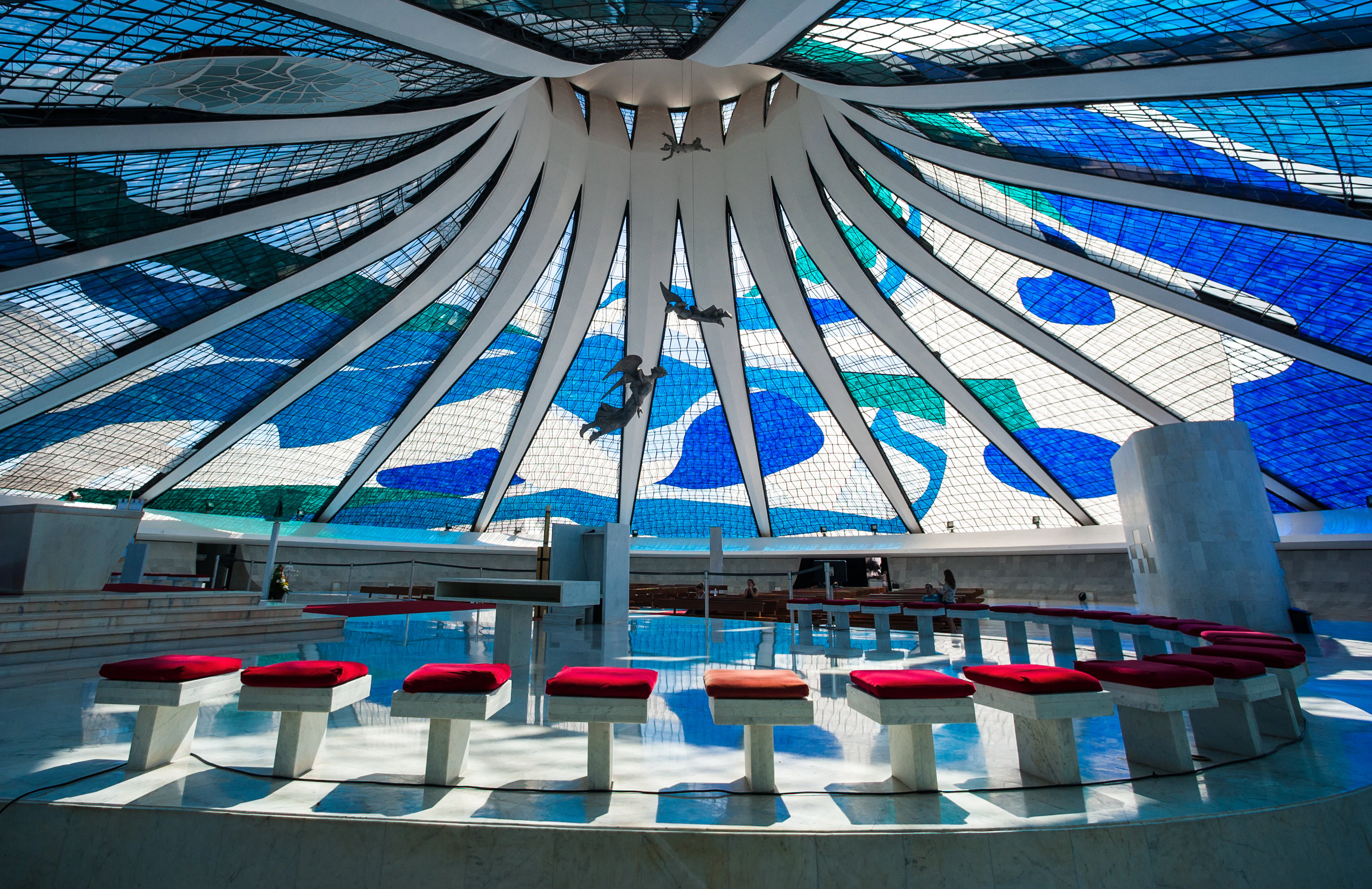 Single benches with red cushions are arranged in a semicircle under a stained glass window with geometric forms.