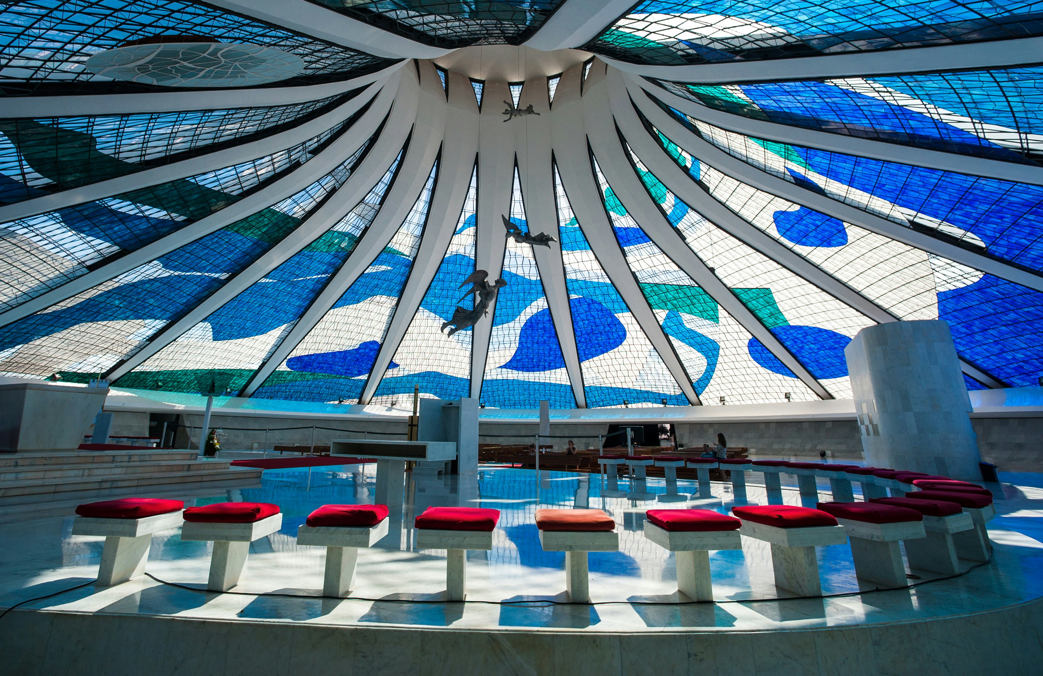 Single benches with red cushions are arranged in a semicircle under a stained glass window with geometric forms.