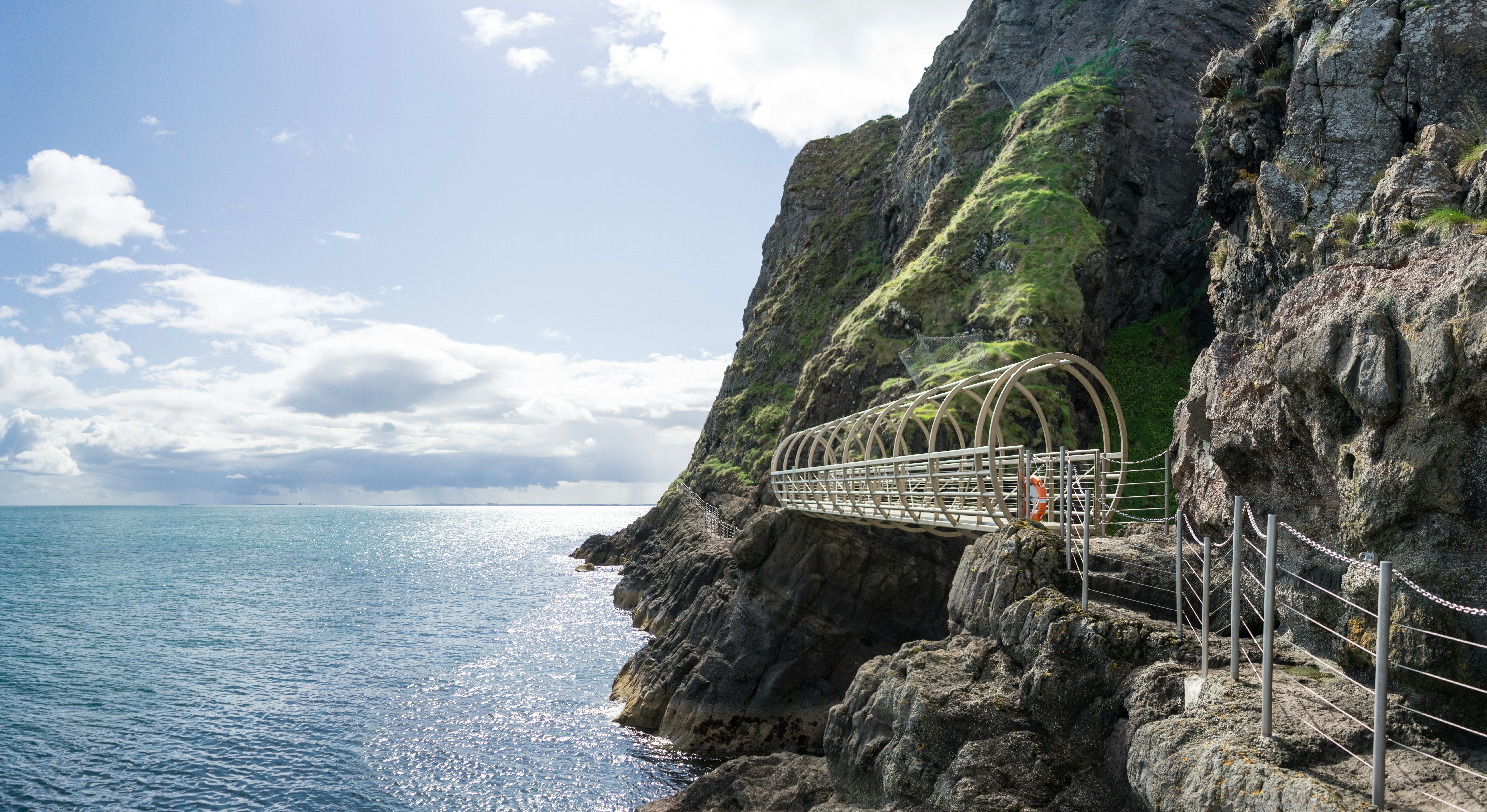 A metal walkway leads along the face of a steep cliff by the sea.