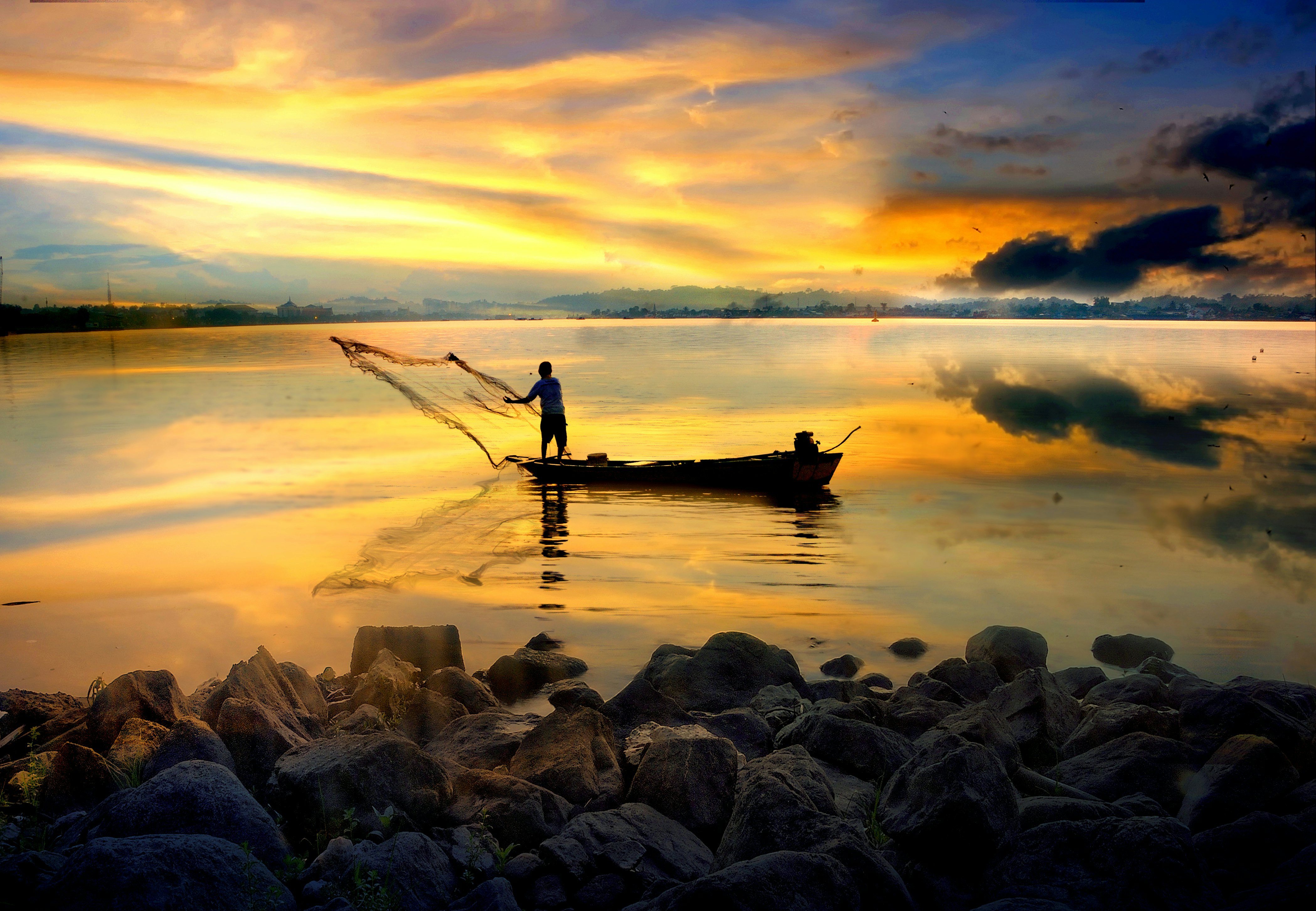 A fisherman casting a net from a small boat in a river is seen in silhouette at sunrise.