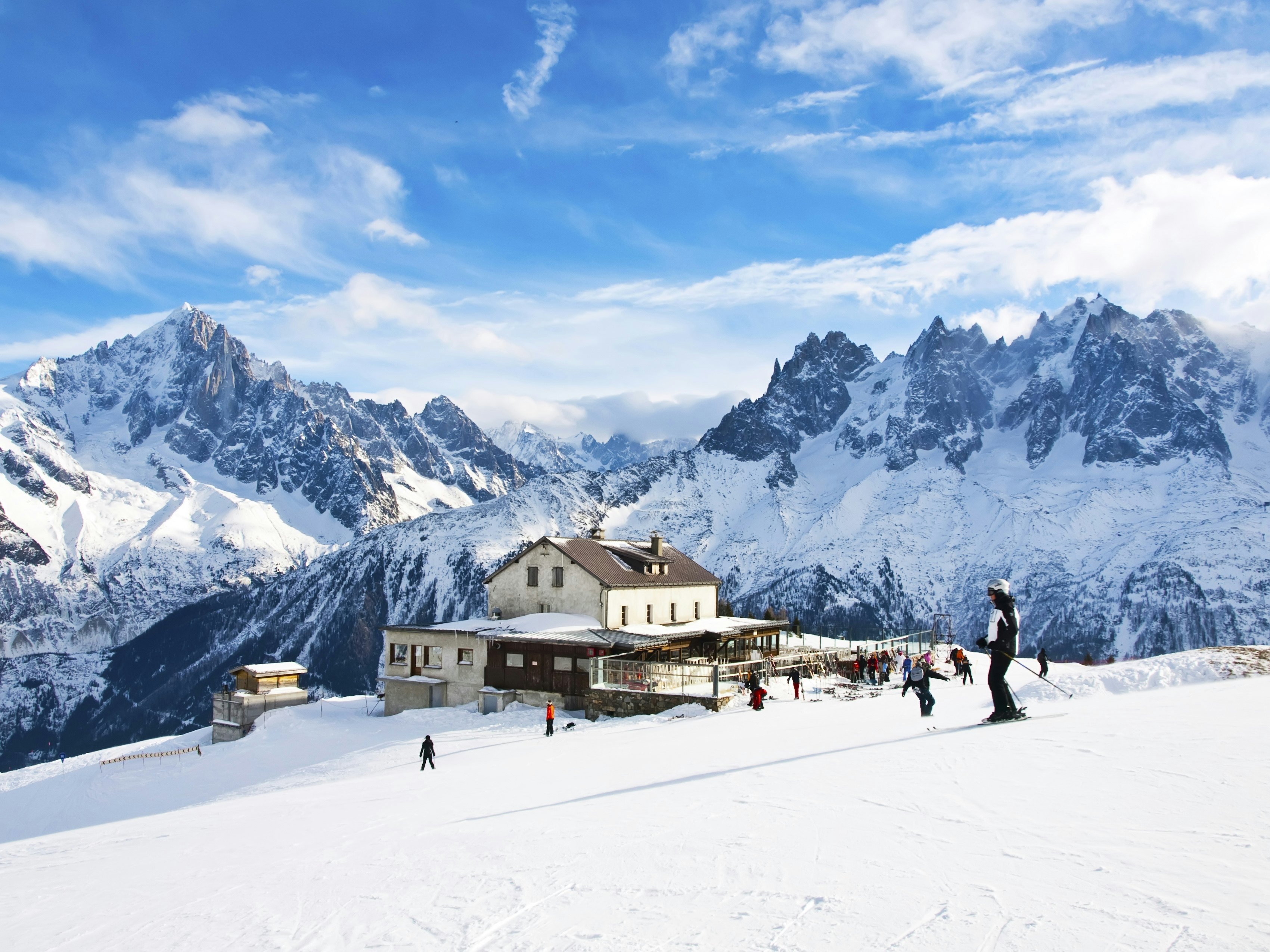 People ski past a lodge on a slope at a resort, with jagged peaks visible in the distance.
