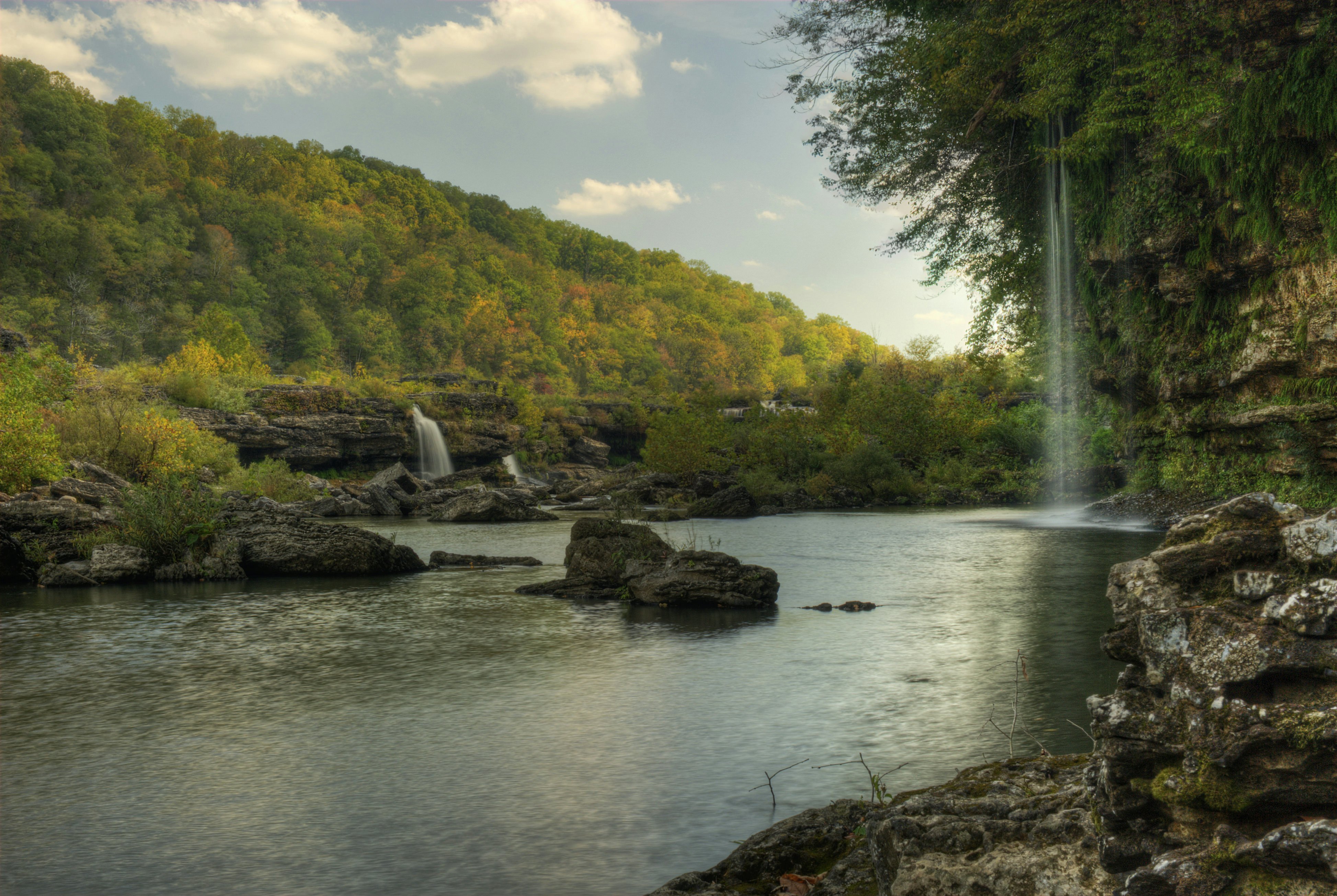 A thin, high waterfall to the right and a lower, denser waterfall in the left distance with a small body of water in front. In the far distance is a forested hillside with just-yellowing leaves.