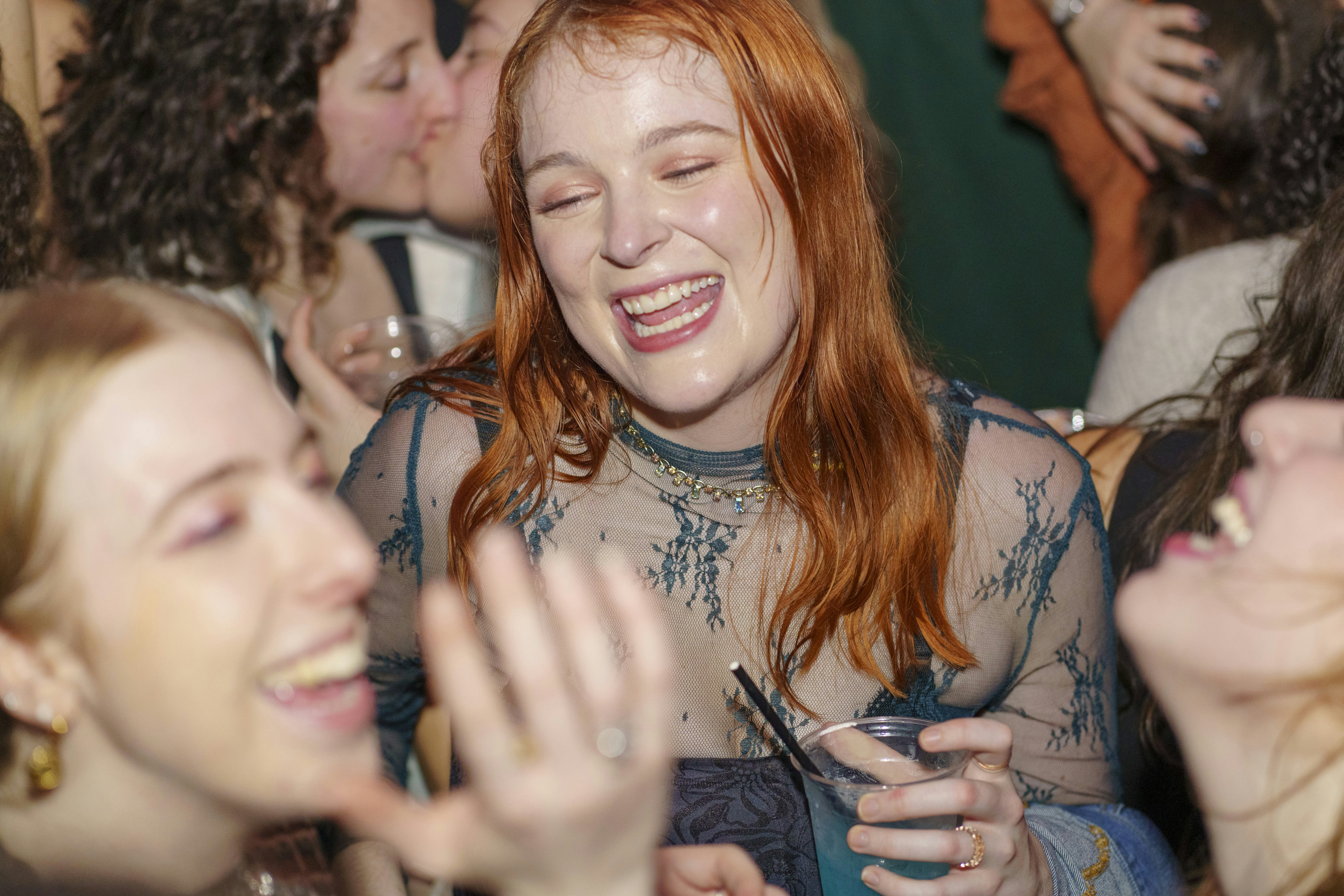 A smiling woman with red hair holds a drink in the center of a crowd.