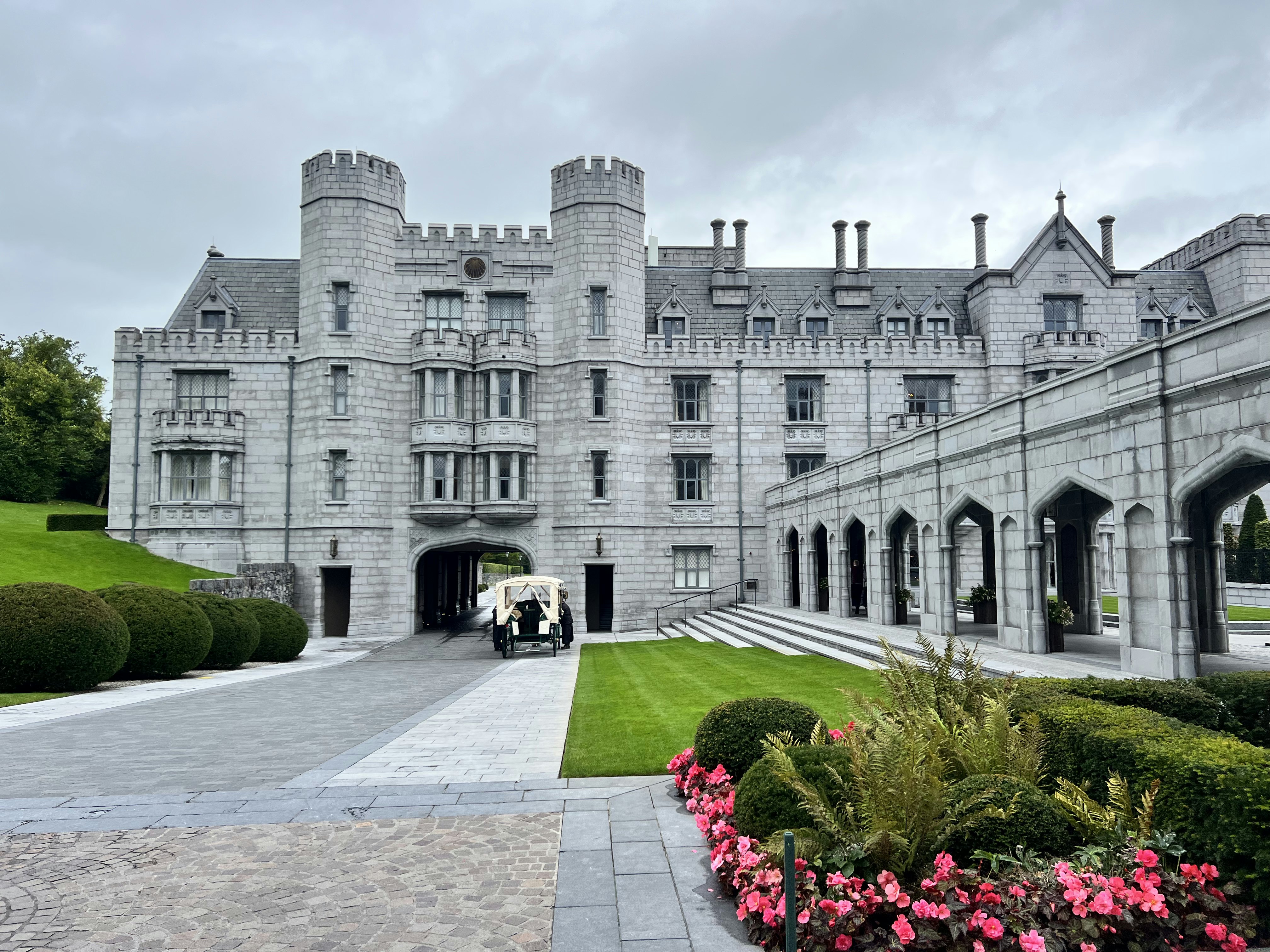 A large gray-stone manor house with pristine garden lawns and flower beds.
