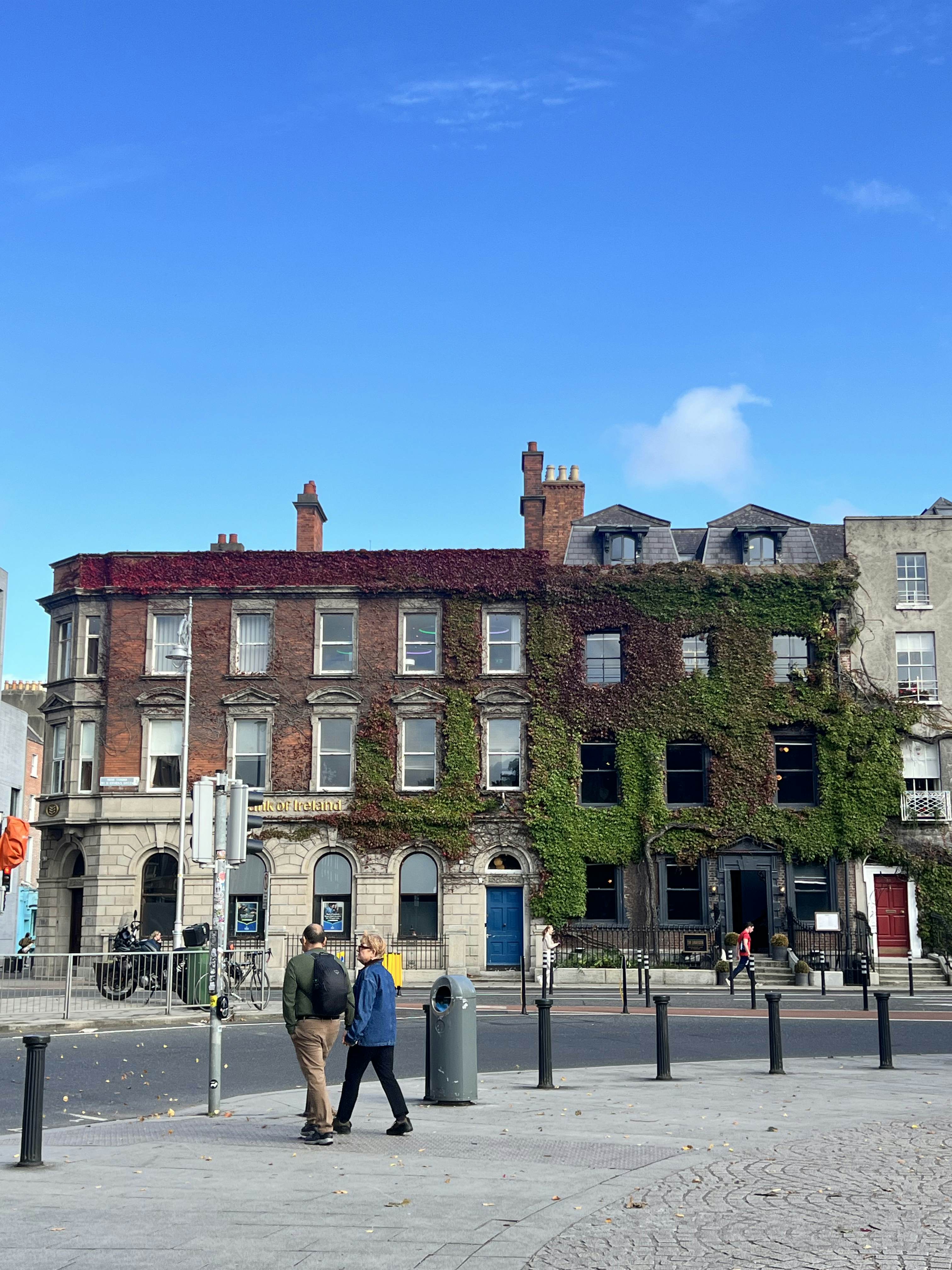 Pedestrians cross a city street in front of buildings covered in ivy.