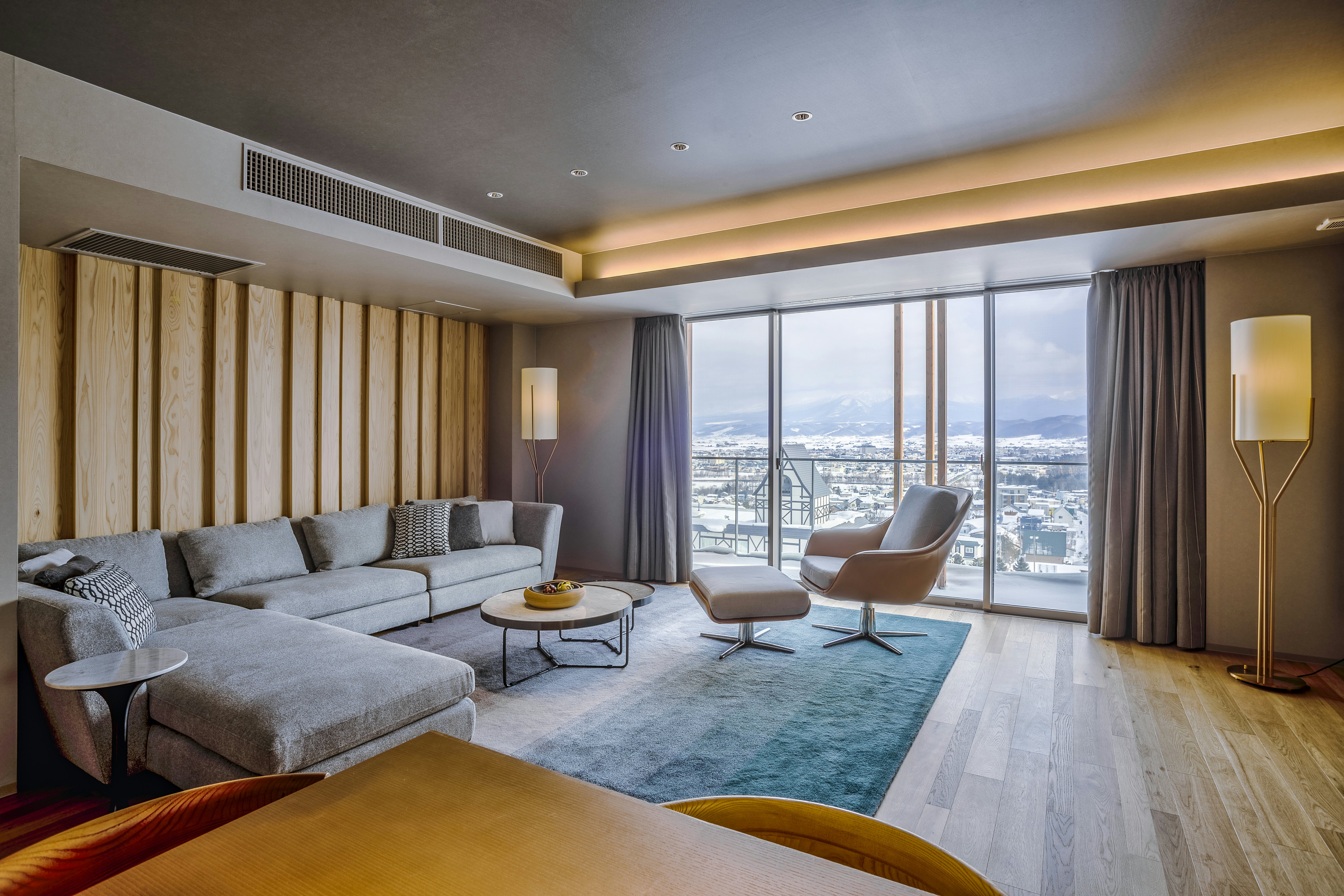A large hotel room with a corner sofa in a lounge area facing a floor-to-ceiling window with city and mountain views.
