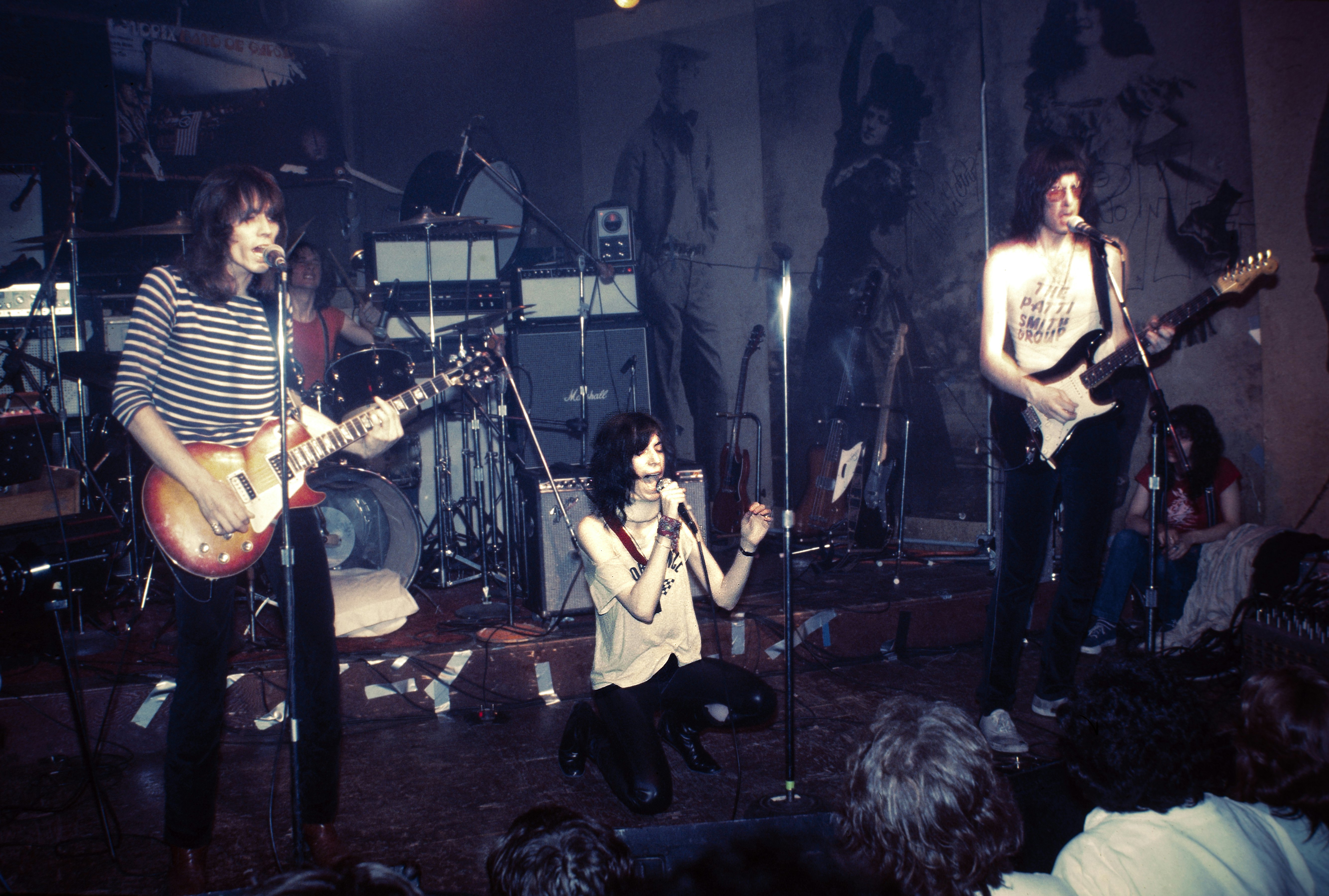 A woman kneels on a stage with a microphone wearing a white tshirt two people flank her playing the guitar a drums and speakers behind her.