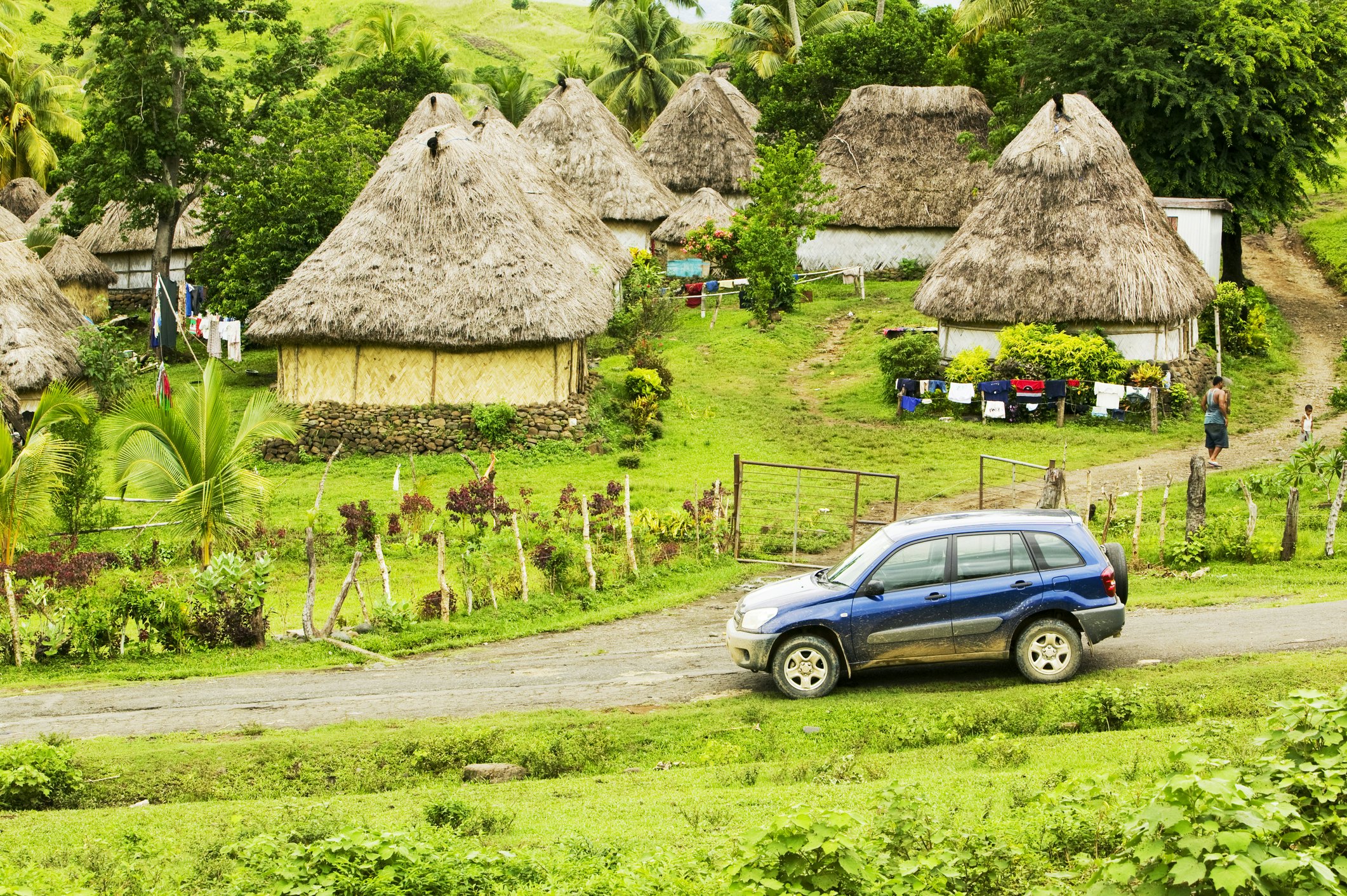 A four-wheel drive car on a dirt track at the edge of a village of thatched huts.