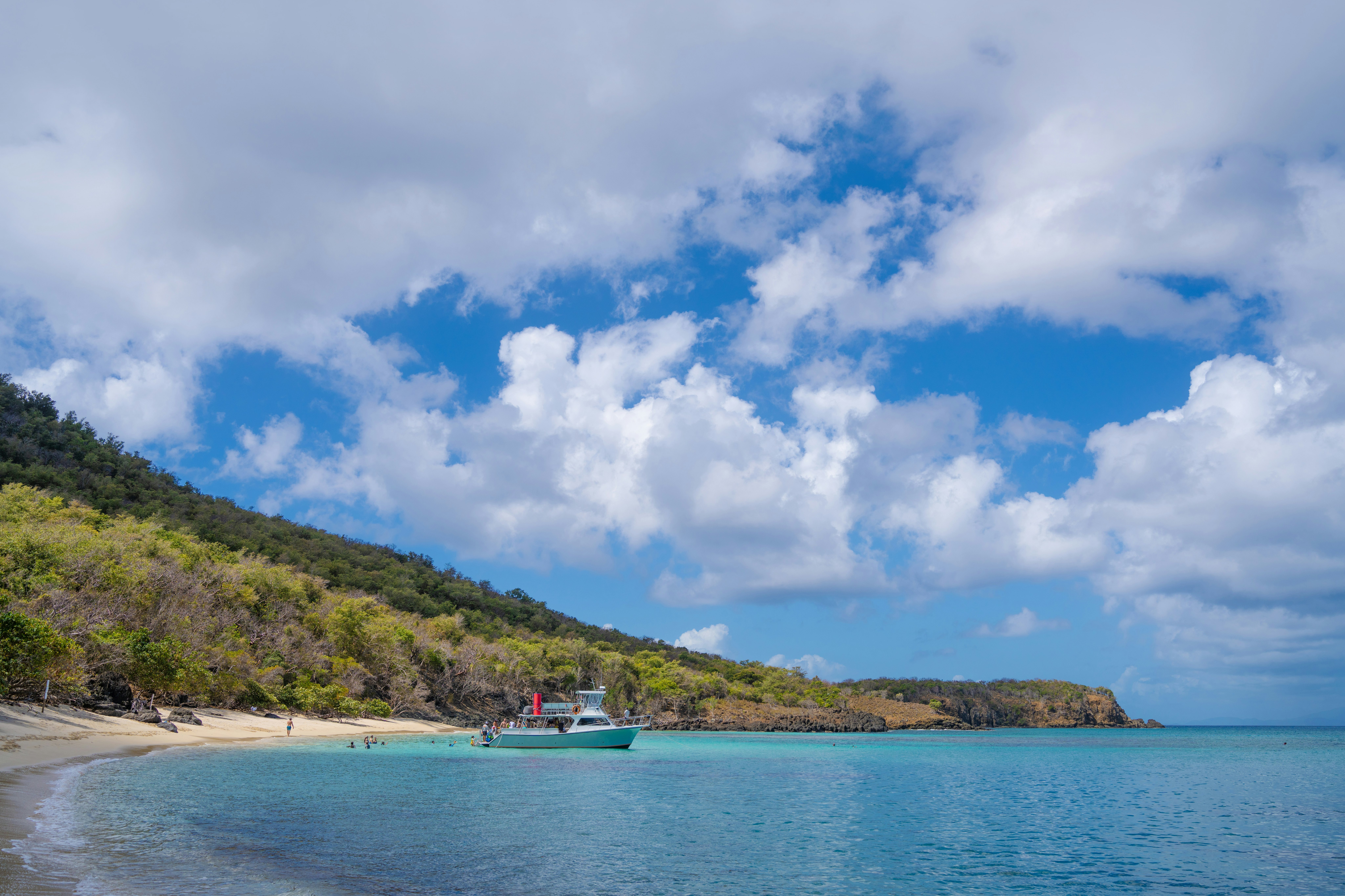 A boat moored in turquoise water by a sandy beach in Puerto Rico; the hillside is covered with green vegetation, and the sky is blue with white clouds.