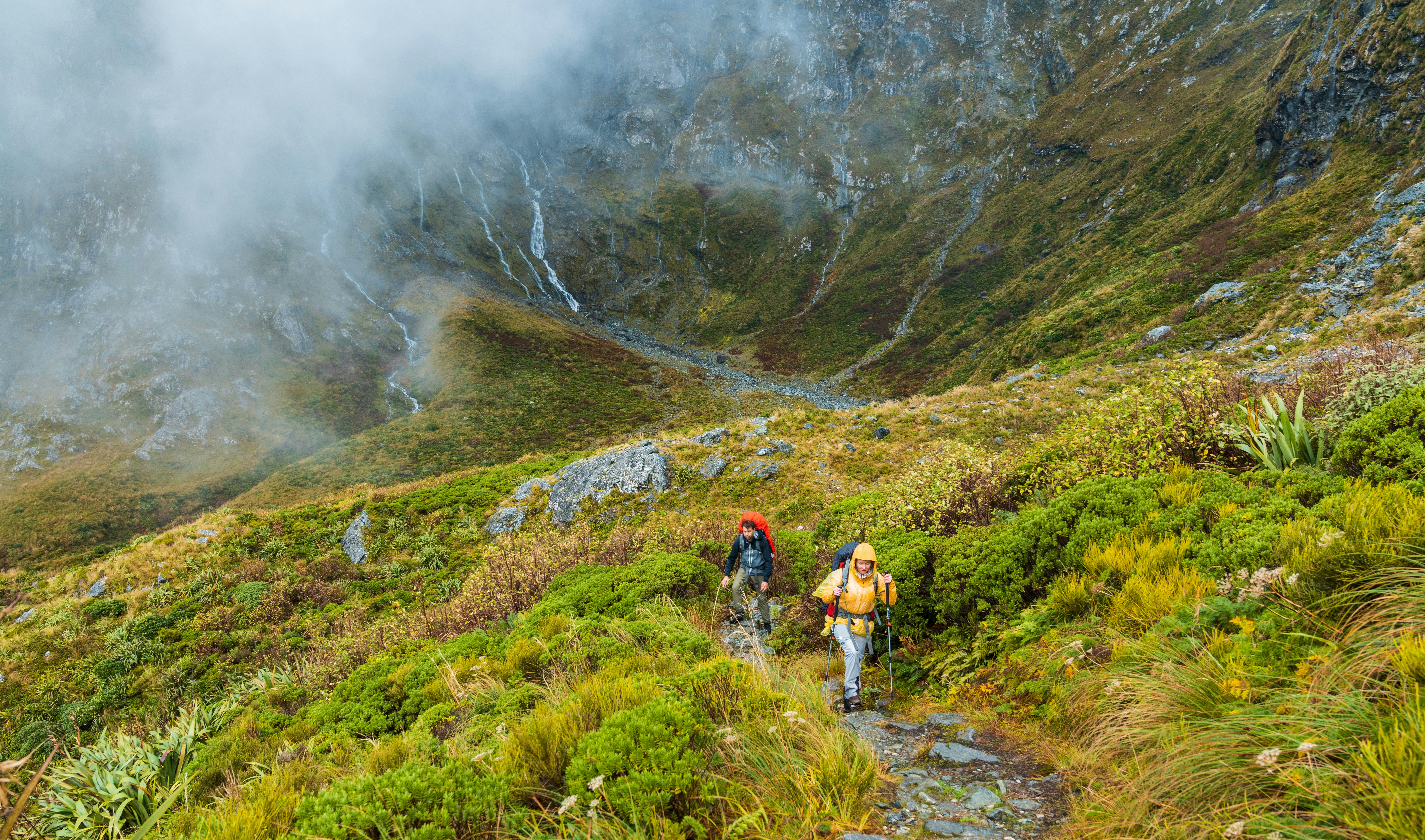 Two hikers carrying gear and walking with poles follow a rocky trail uphill on a misty damp day.