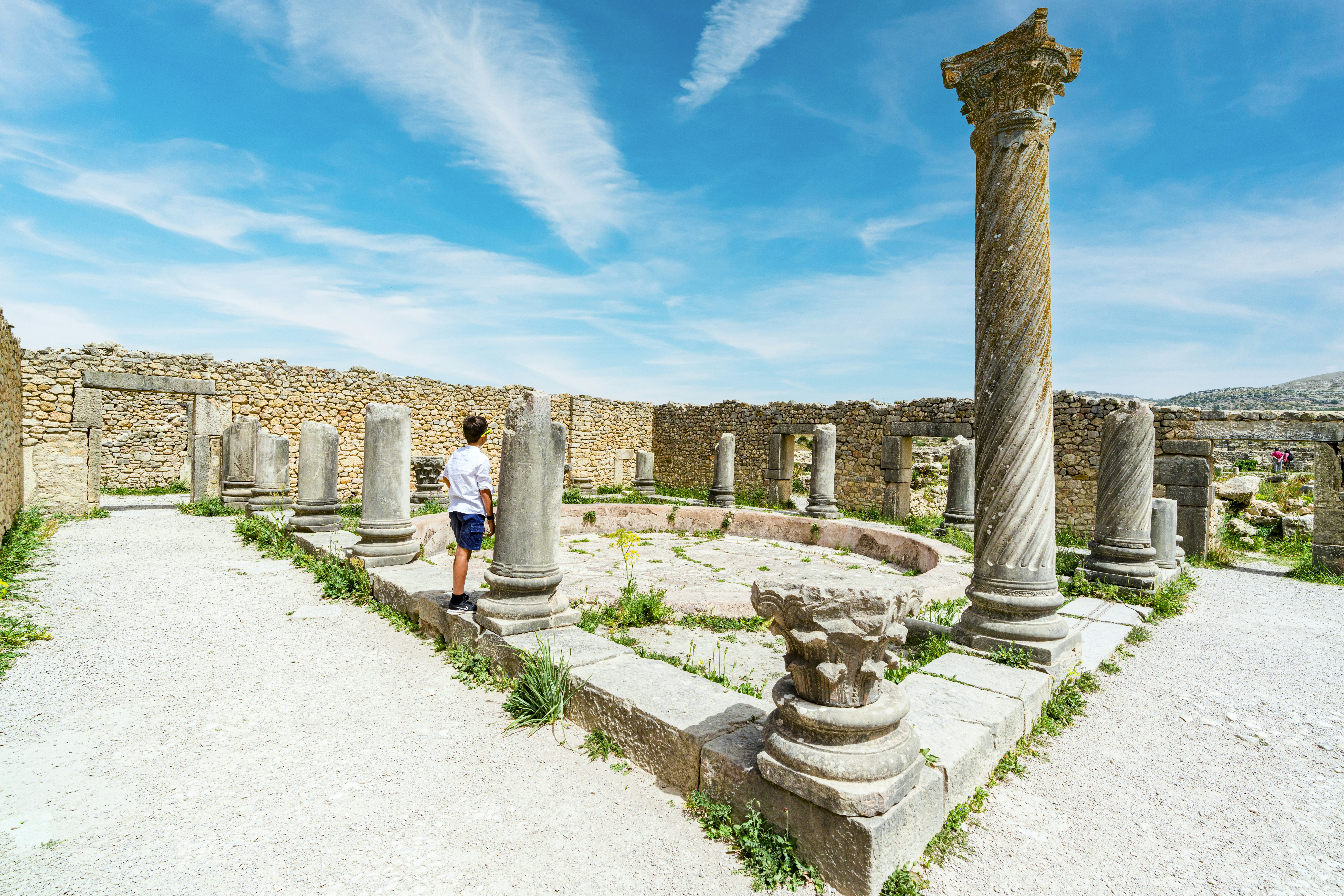 A young boy aged around 10 years old stands near an ancient Roman column looking at an archaeological site.
