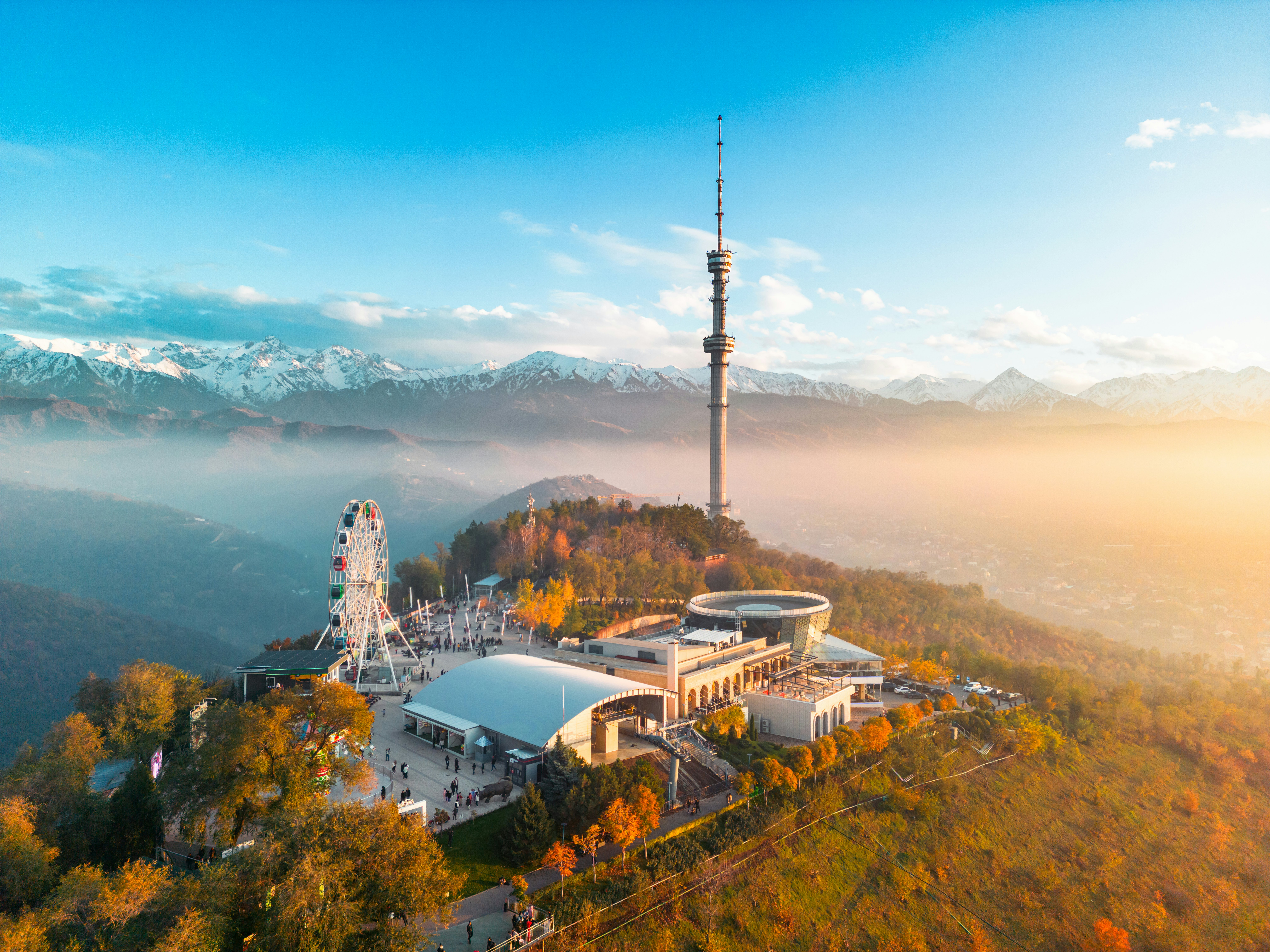 An aerial view of an amusement park and television tower at the top of a hill, shrouded in mist. Snow-capped mountains are seen in the distance.