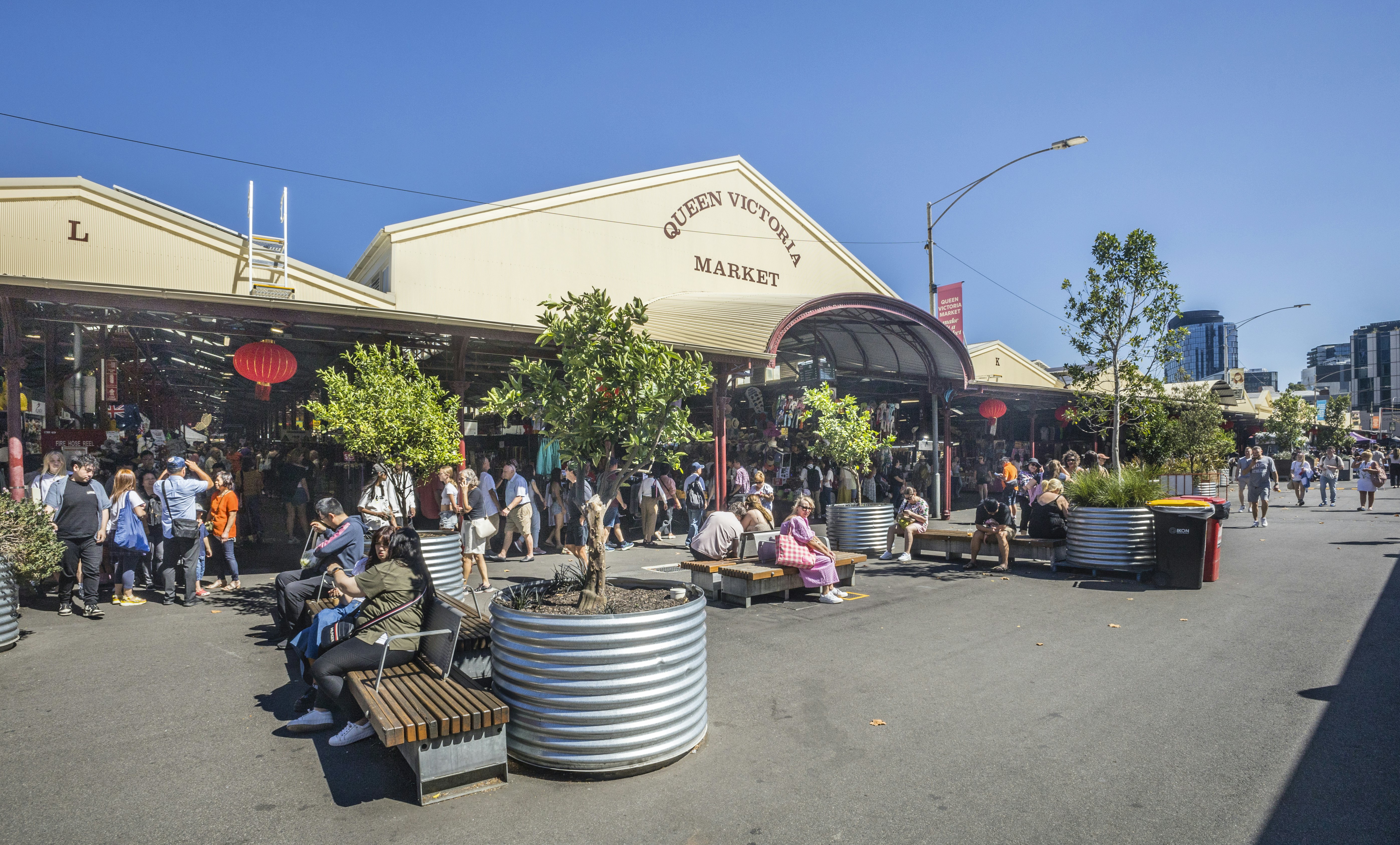 People sit on benches in the sunshine outside a large covered market place.
