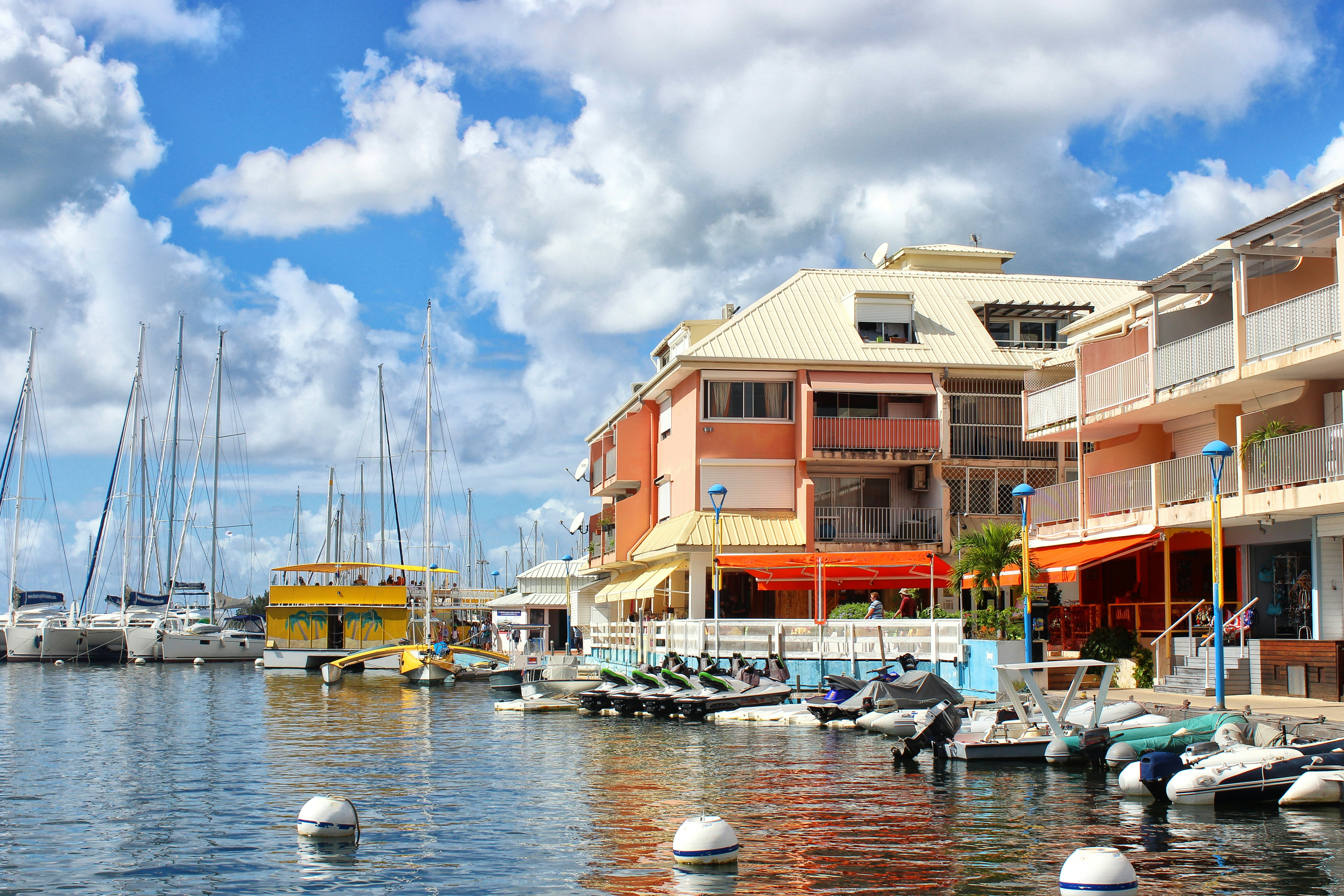 Boats and Jet Skis docked at a harbor with a colorful peach-orange building bordering a walkway along the water on a sunny day.