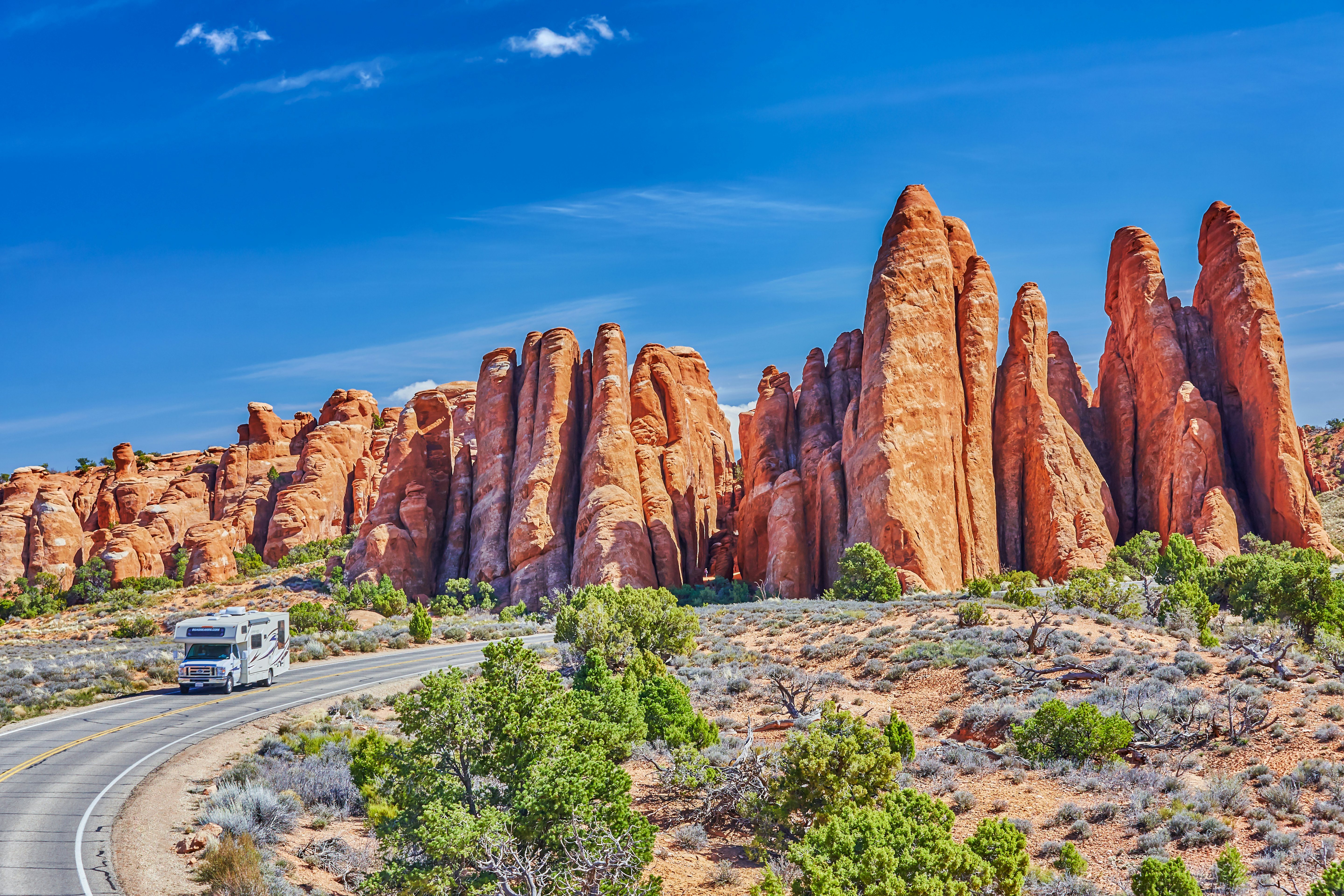 An RV drives on a two-lane blacktop road around a curve with reddish rock formations behind it; the sky is blue, and there are green bushes to the side of the road.