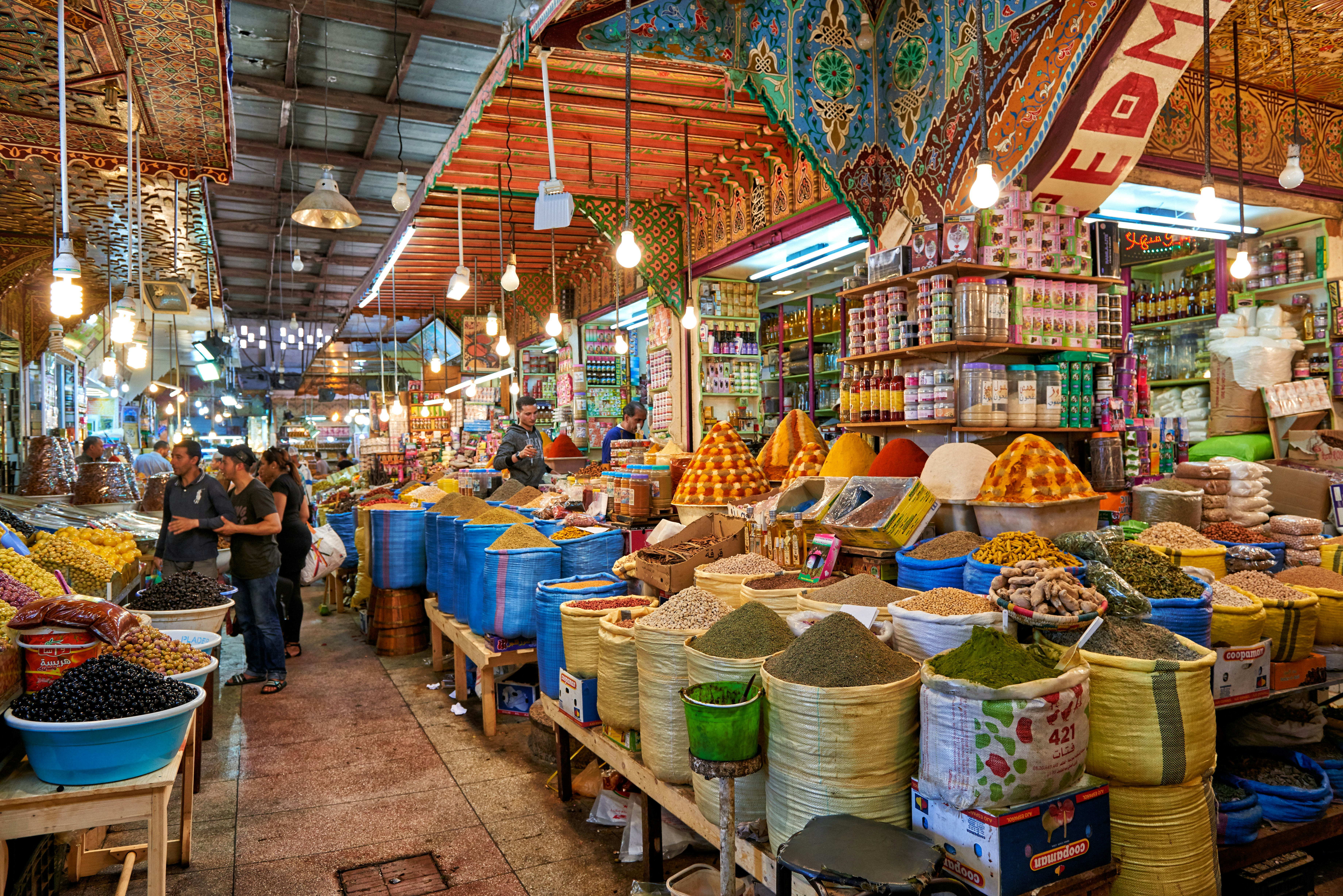 A covered market place with stalls selling spices and olives piled high in bags and baskets.