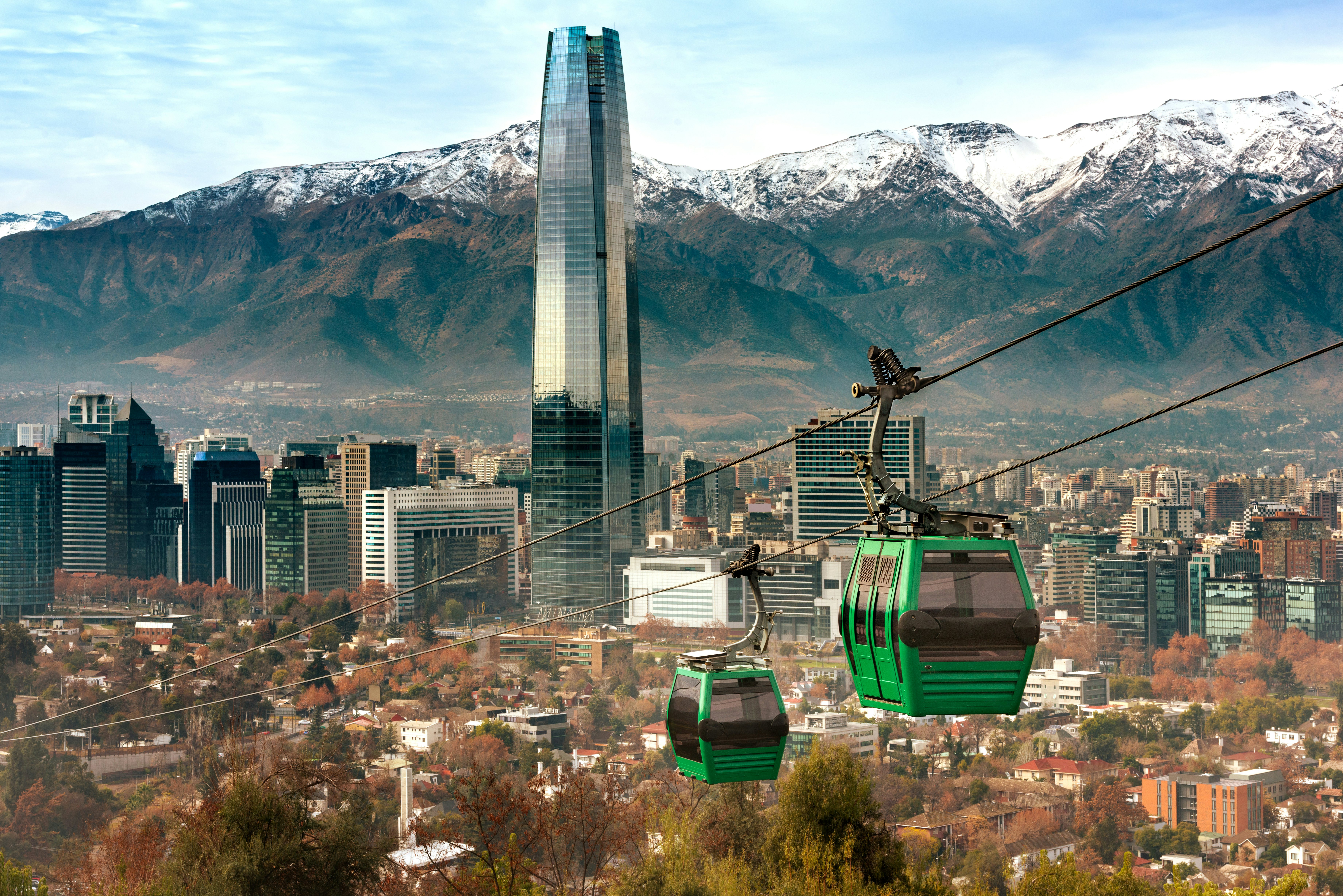 Green gondolas ascend a hill on a cable, with a view of a city skyline and snow-topped mountains in the distance.