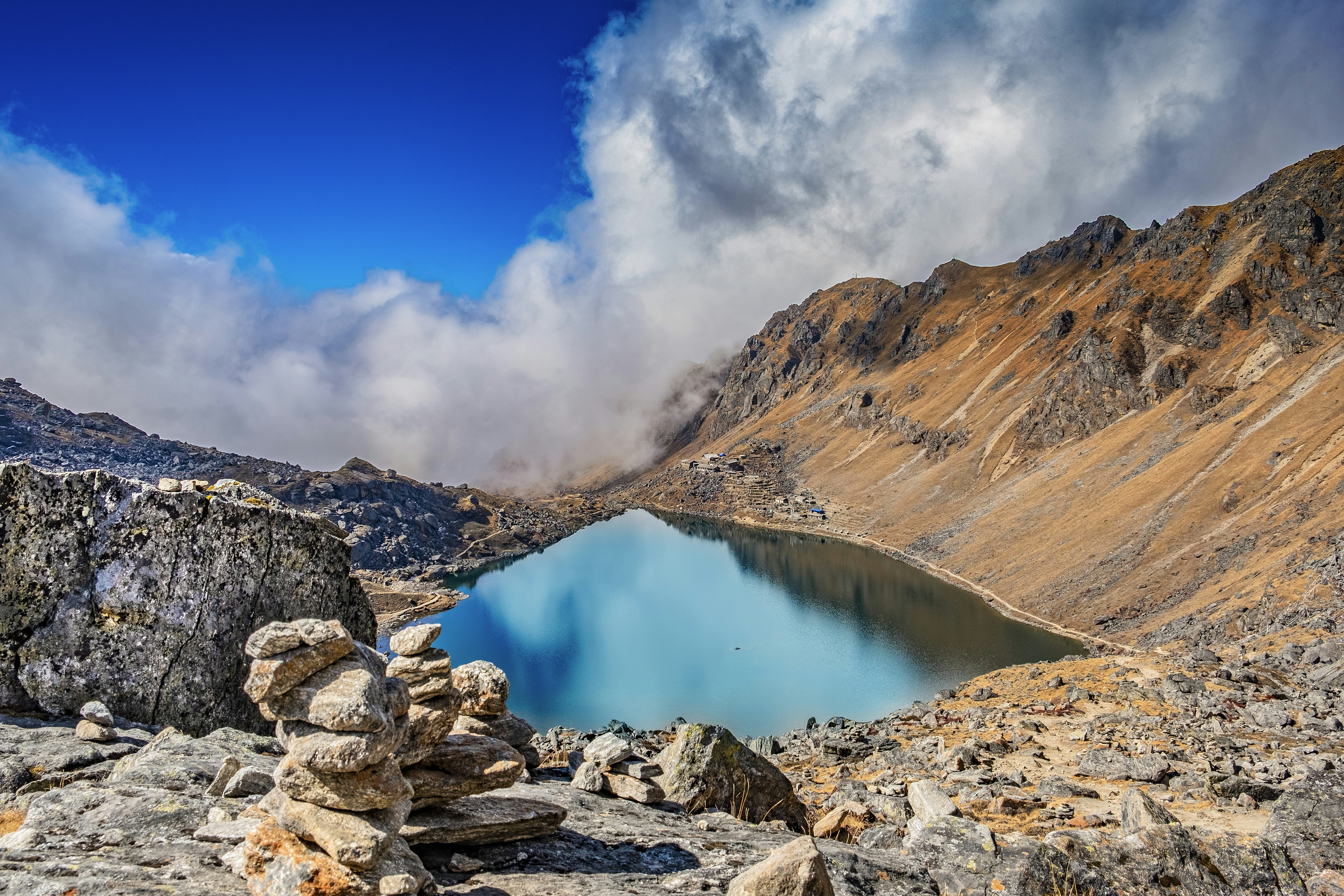 A view from above of a glassy, high-altitude lake. Rocky slopes lead to its shores, with clouds hanging low in the sky.