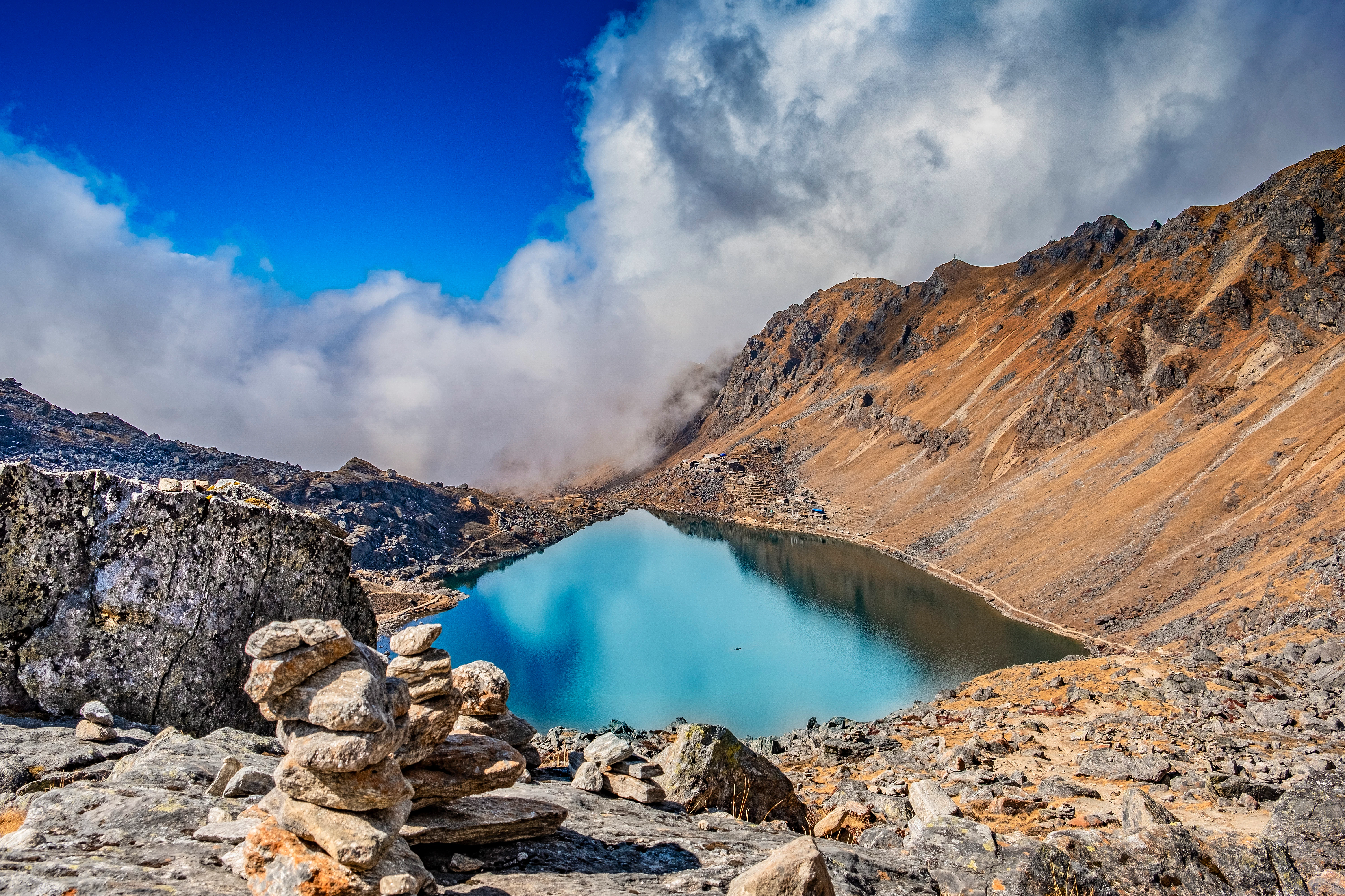 A view from above of a glassy, high-altitude lake. Rocky slopes lead to its shores, with clouds hanging low in the sky.