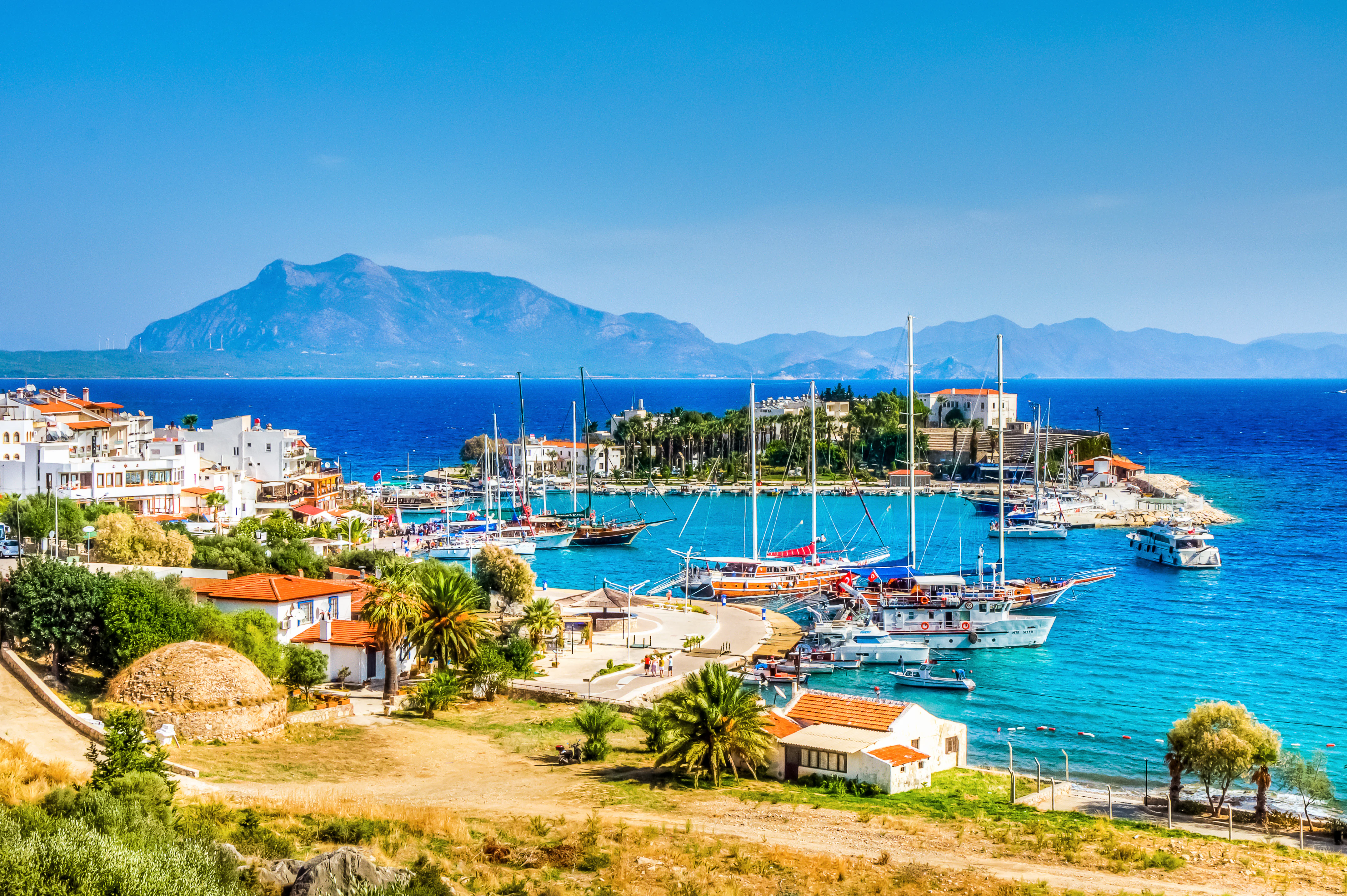 Boats in a harbor of azure water; there is a small town of white buildings on the shore and mountains in the distance.