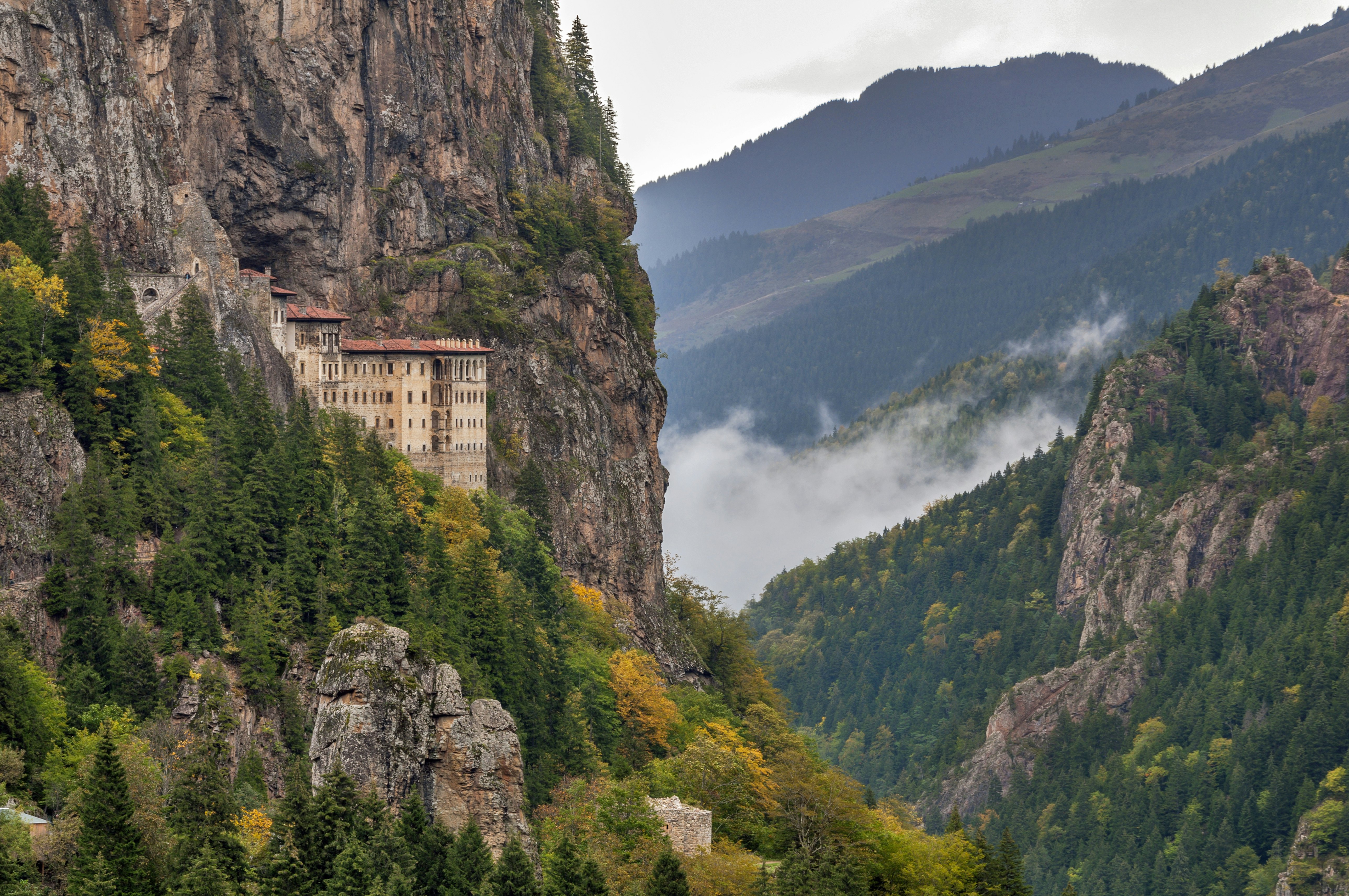 A building sits on the edge of a sheer rocky cliff in Türkiye; there is a cloud in a deep valley in the background.