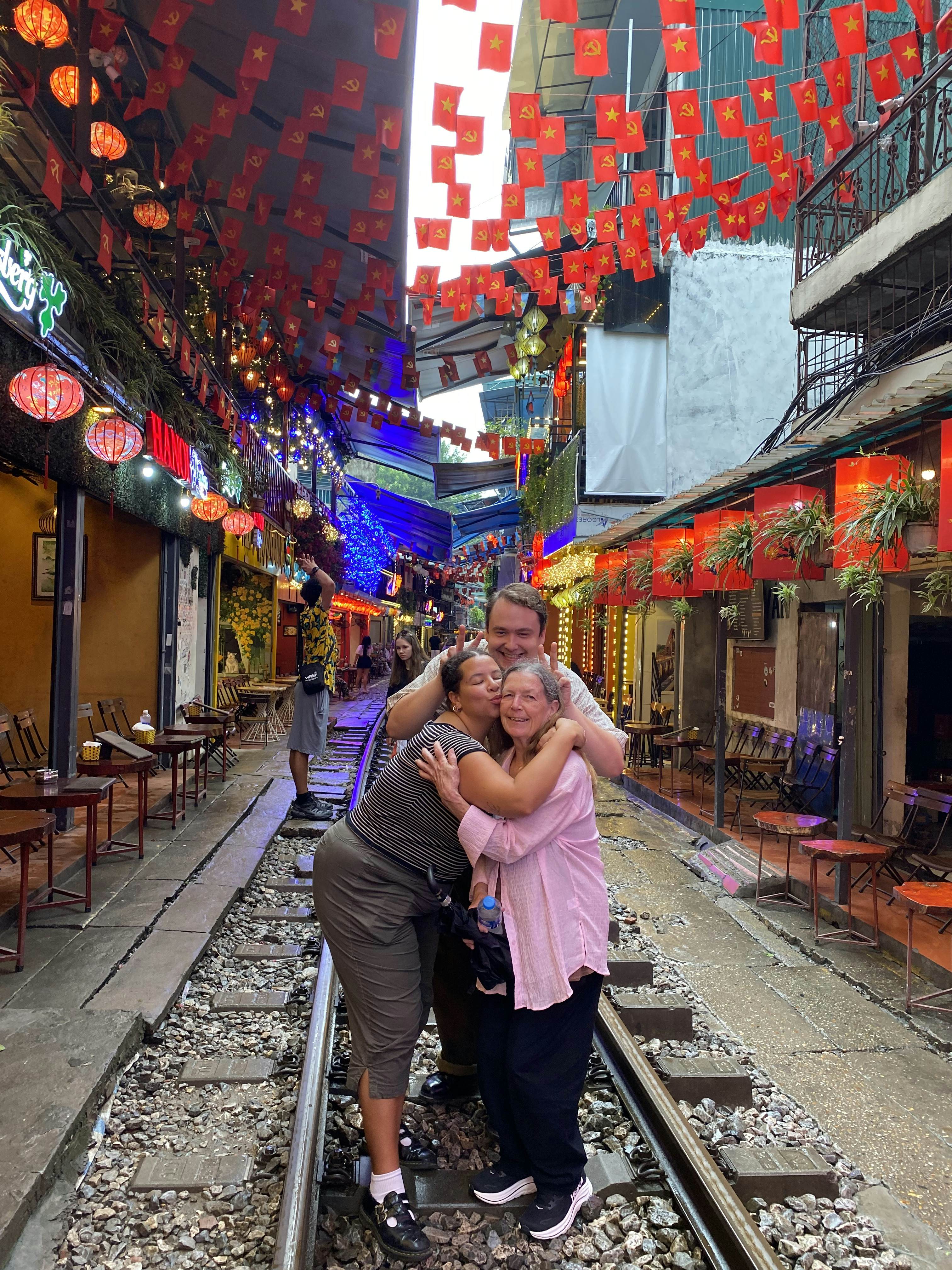 Two women and a man with their arms around each other, smiling while standing on a train track