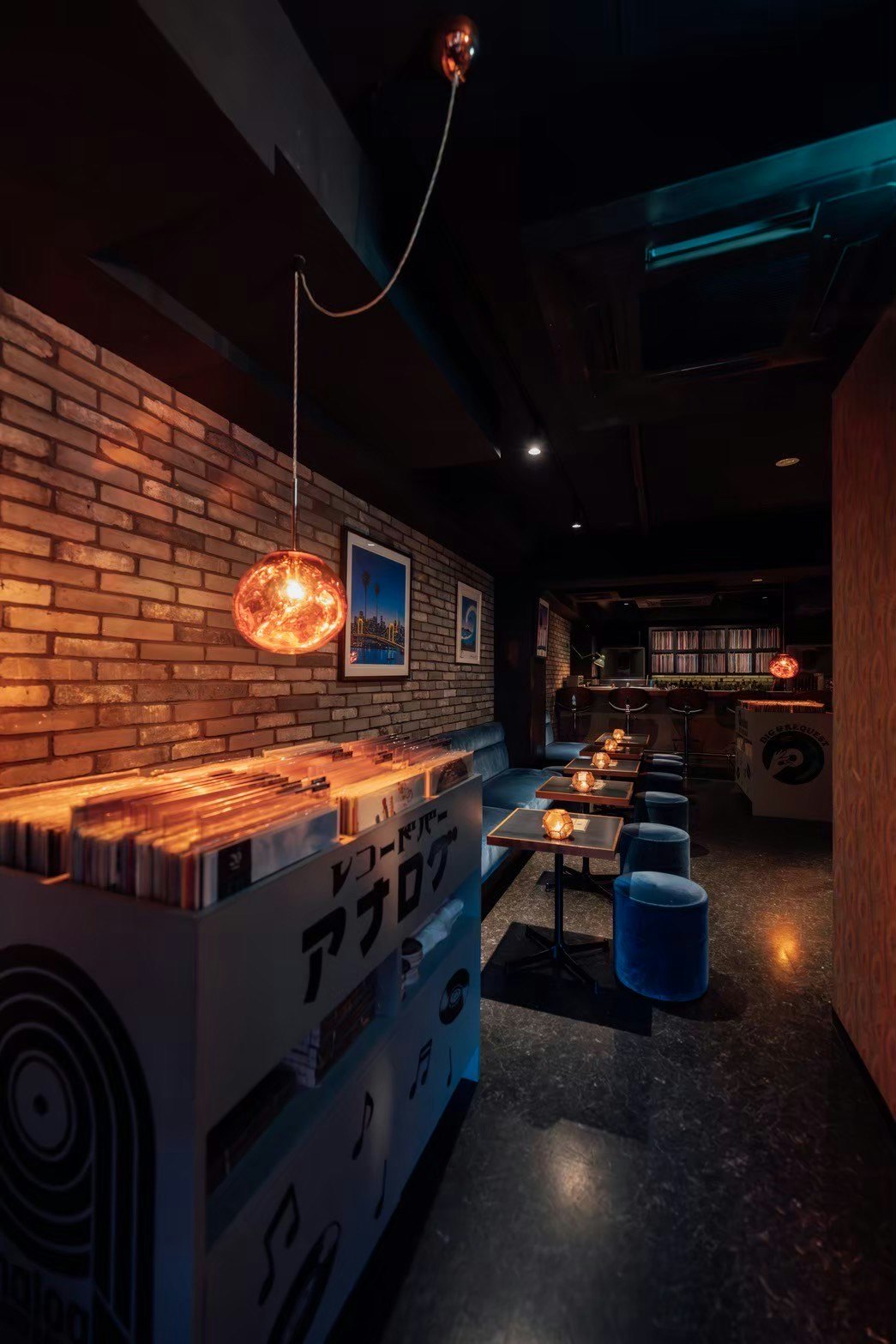 A box of records stands in front of a short row of seating and tables. A bar at the back of the room is backed by a vast collection of records.