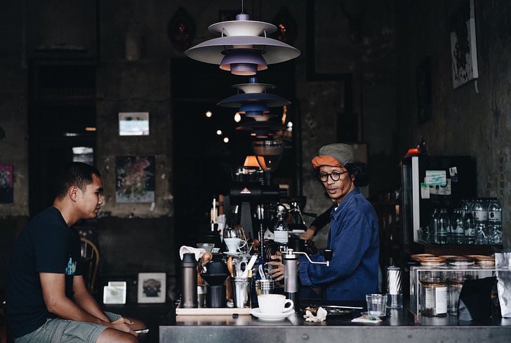 A barista preparing a drink behind a counter in a darkened coffeehouse while a patron waits on the other side