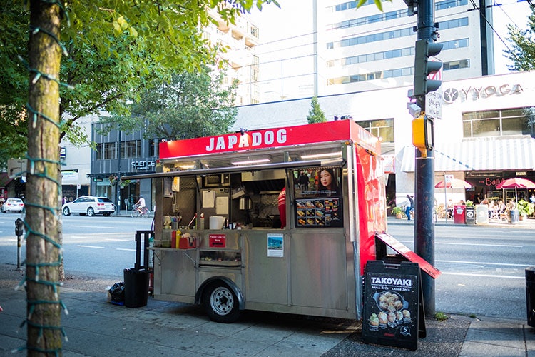 A small hot-dog stand on the sidewalk of a city street with a large sign that reads "Japadog".