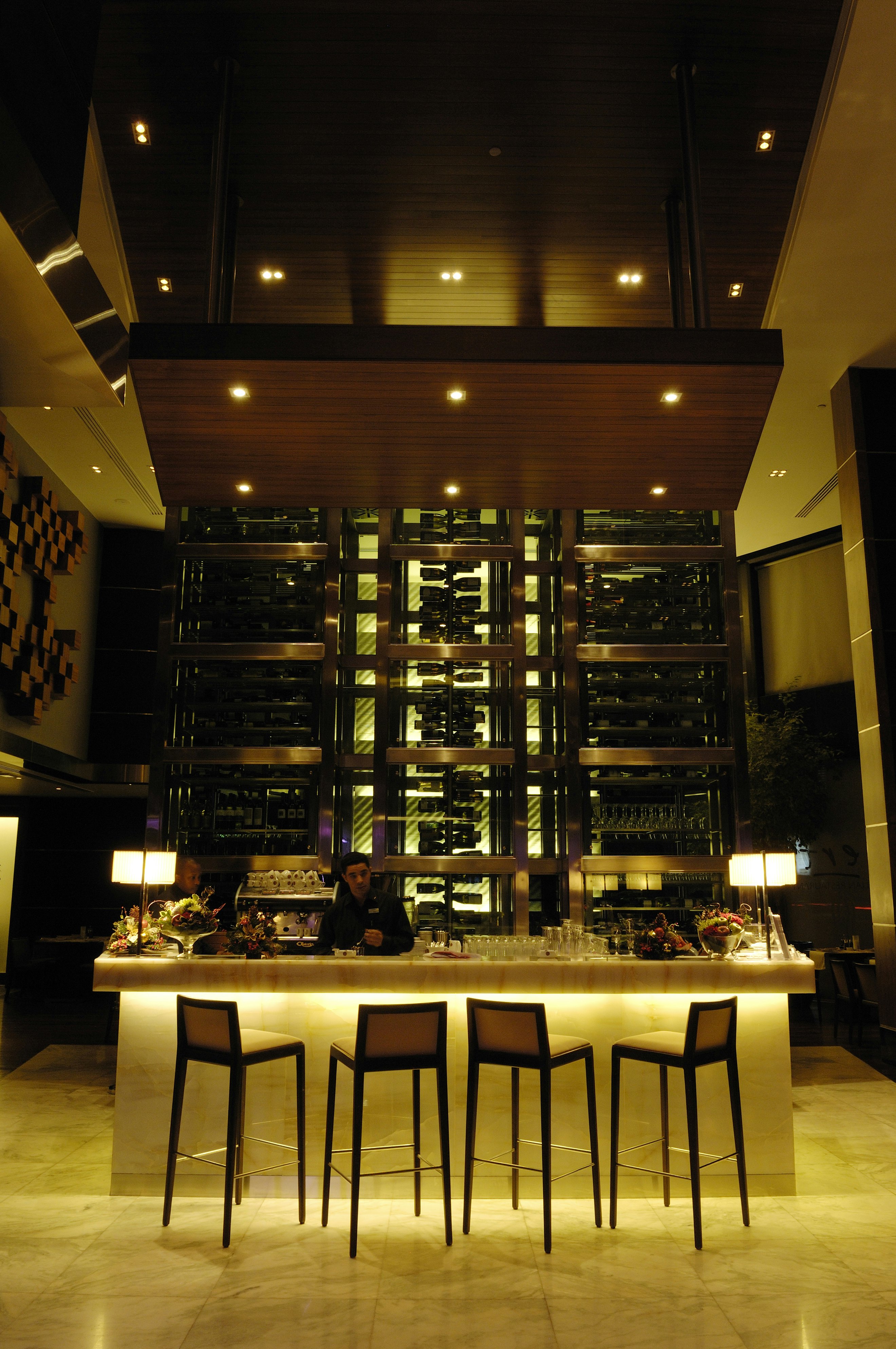 A person stands behind a bar that's lit up underneath, with four tall chairs; bottles are displayed behind the bar.