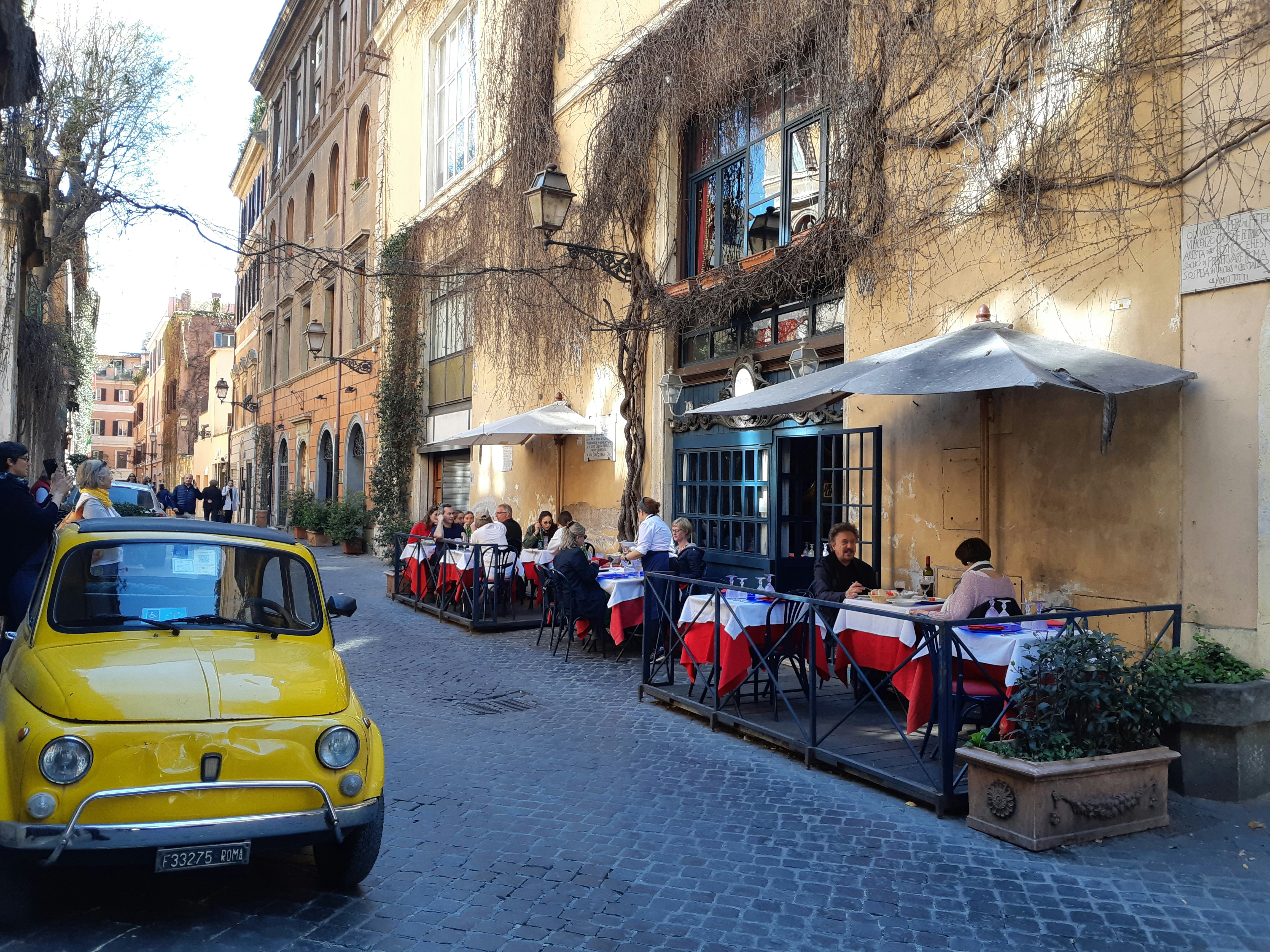 People dining in the street in Rome, with a classic Italian car on the roadside.