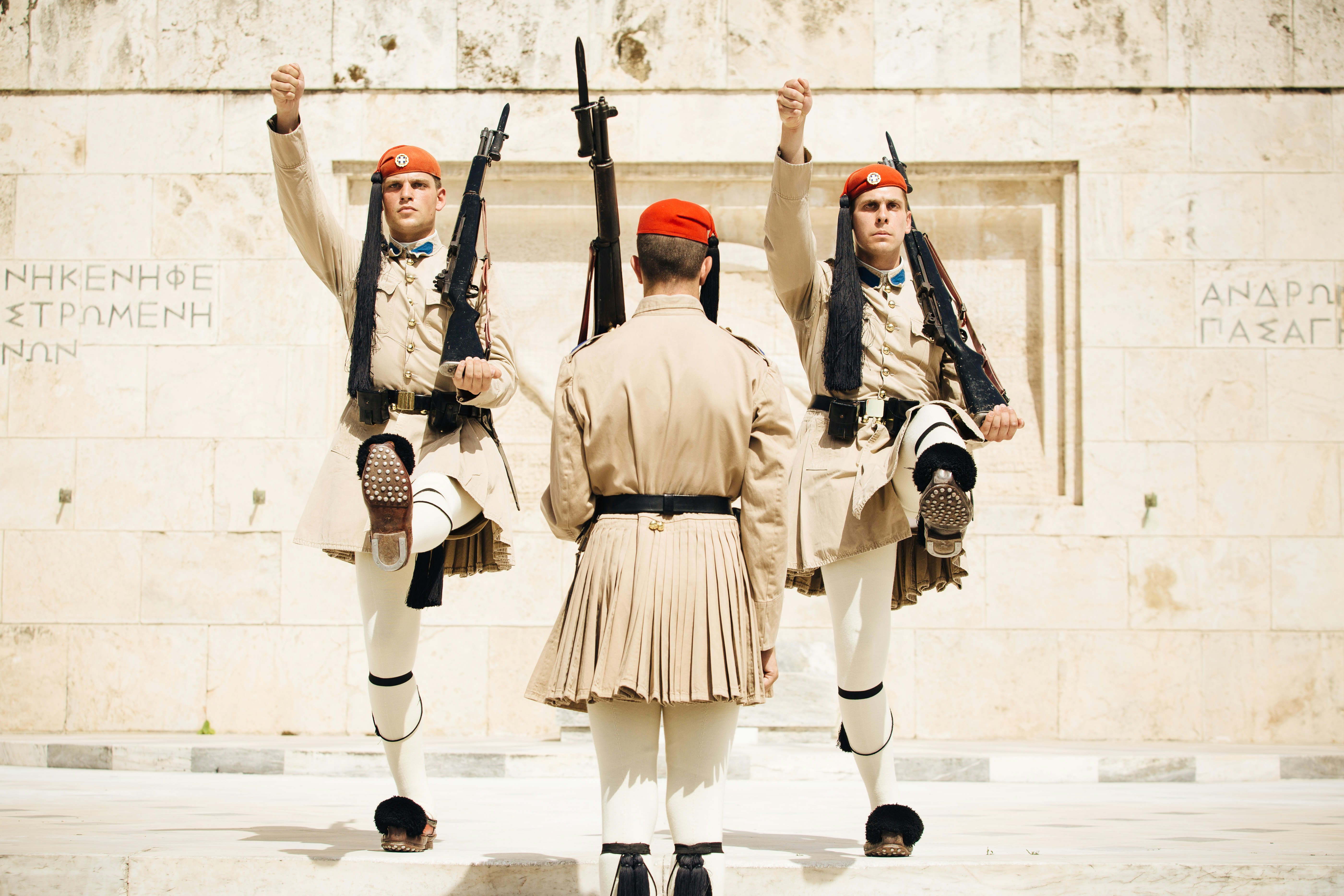 A guard wearing an earth-toned tunic and red beret and holding a bayonet in his left hand has his back toward the camera. Two identically dressed guards face him. Their right arms and left legs are raised, and they also carry bayonets in their left hands.