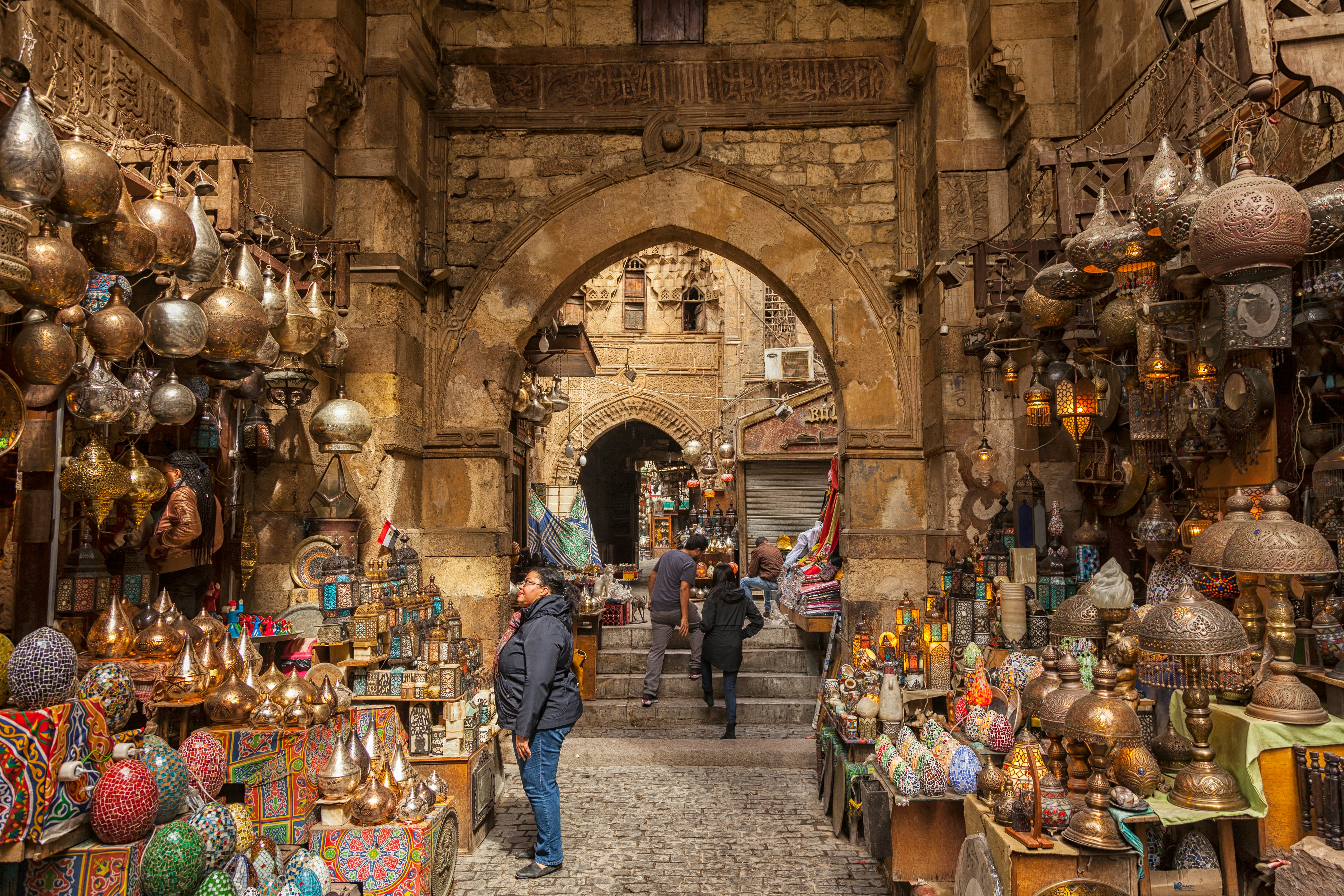 A shopper looks at lanterns hanging from the wall and stacked on shelves at an outdoor stand by an archway on a street in Cairo, Egypt.