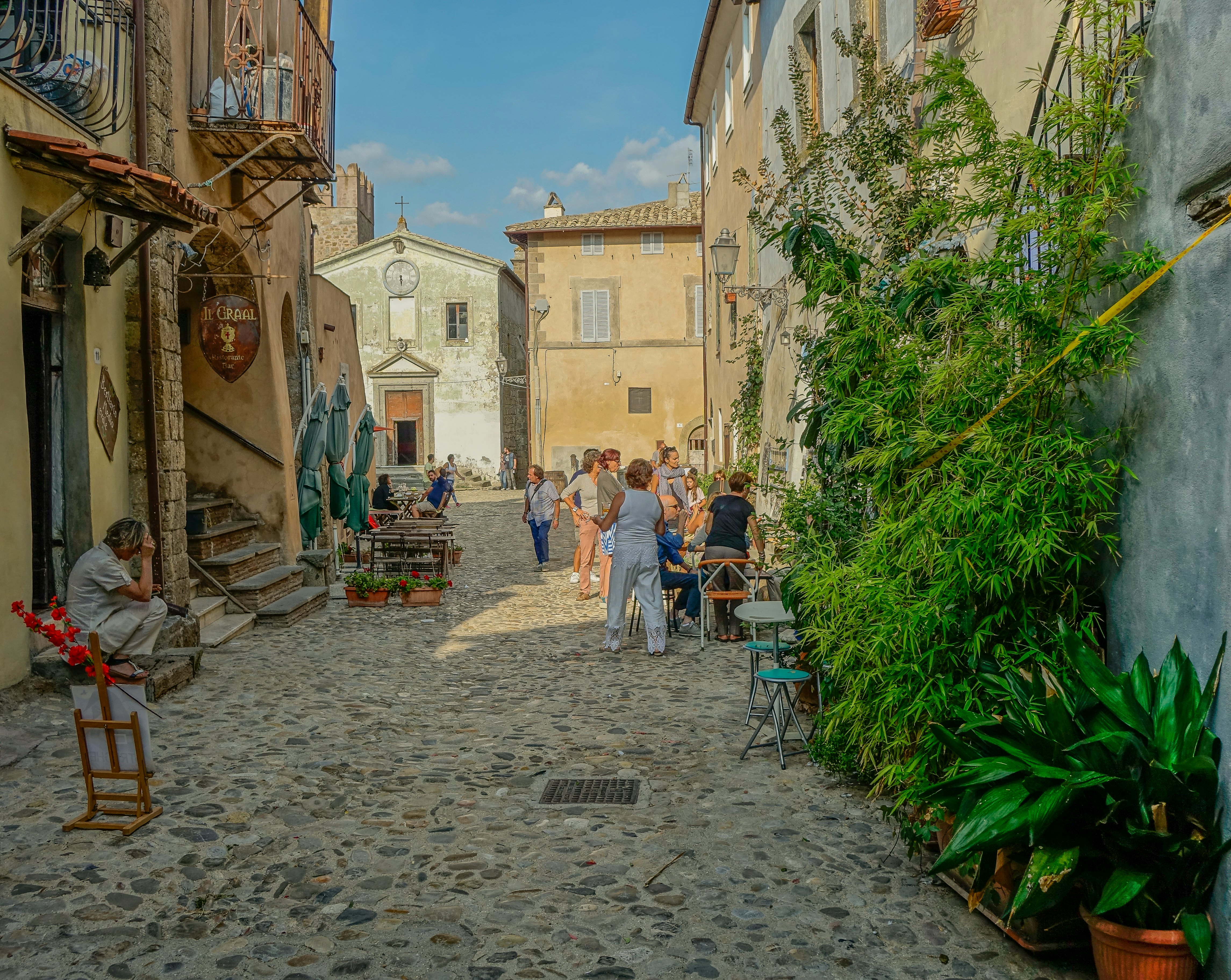 A cobblestone street in a medieval village. People are sitting at tables outside restaurants. There is a church in the distance.